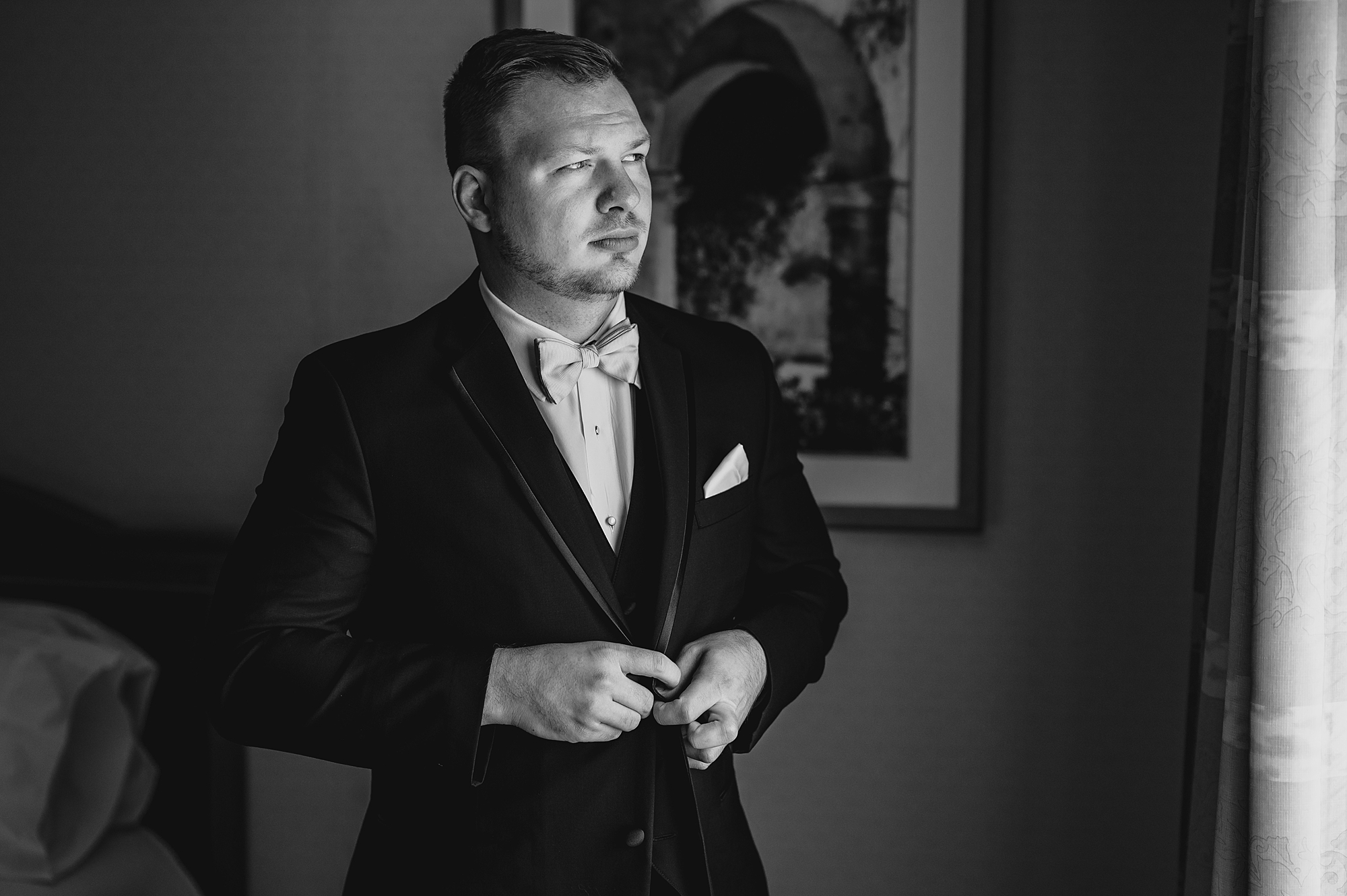 groom adjusts buttons on suit jacket in window at wedding at the Garrison