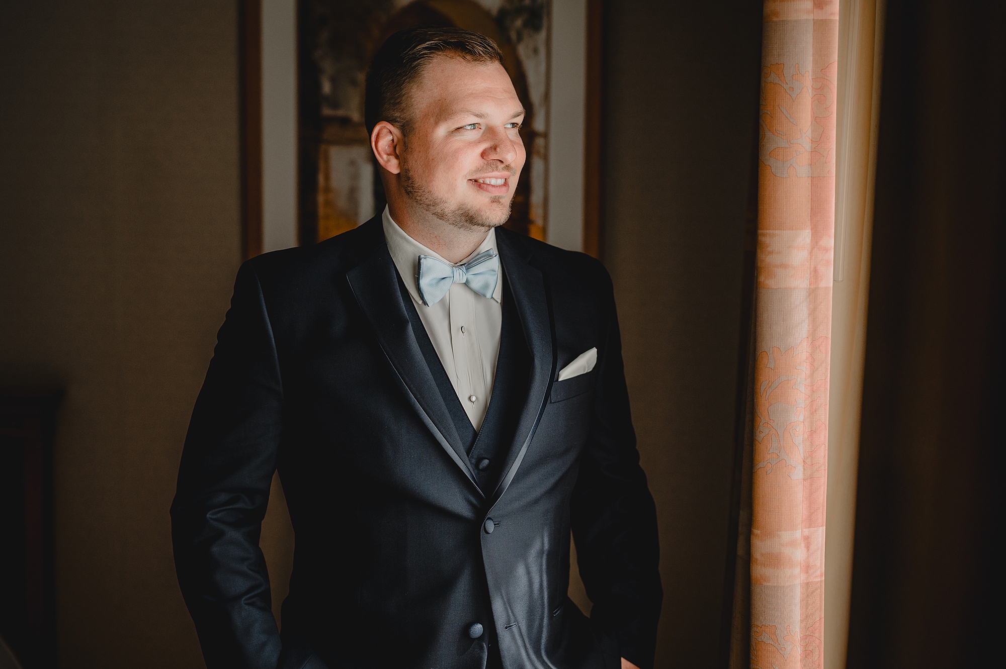 groom leans against window in black suit with blue tie