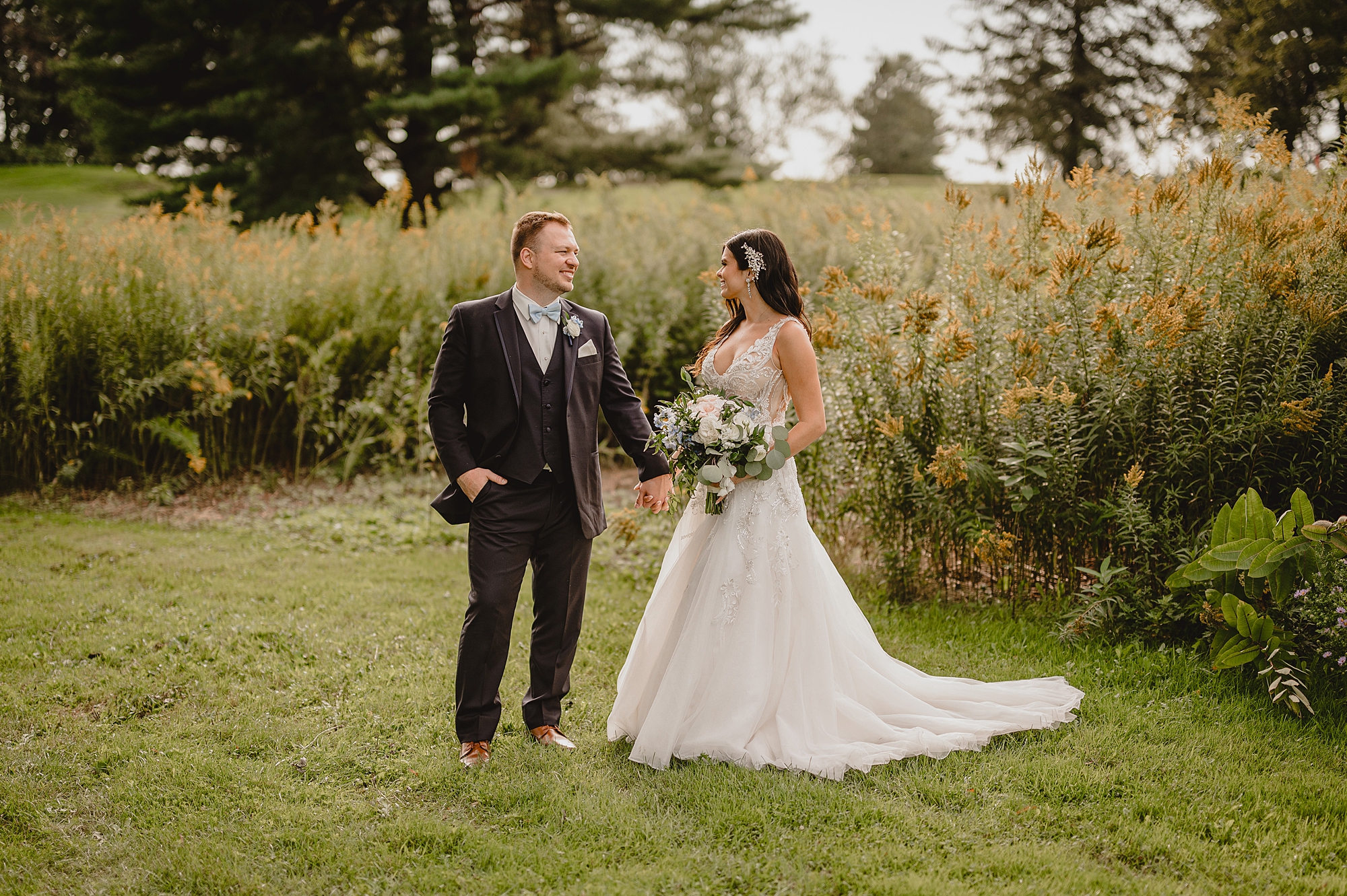 bride and groom hold hands by tall grass on property at the Garrison