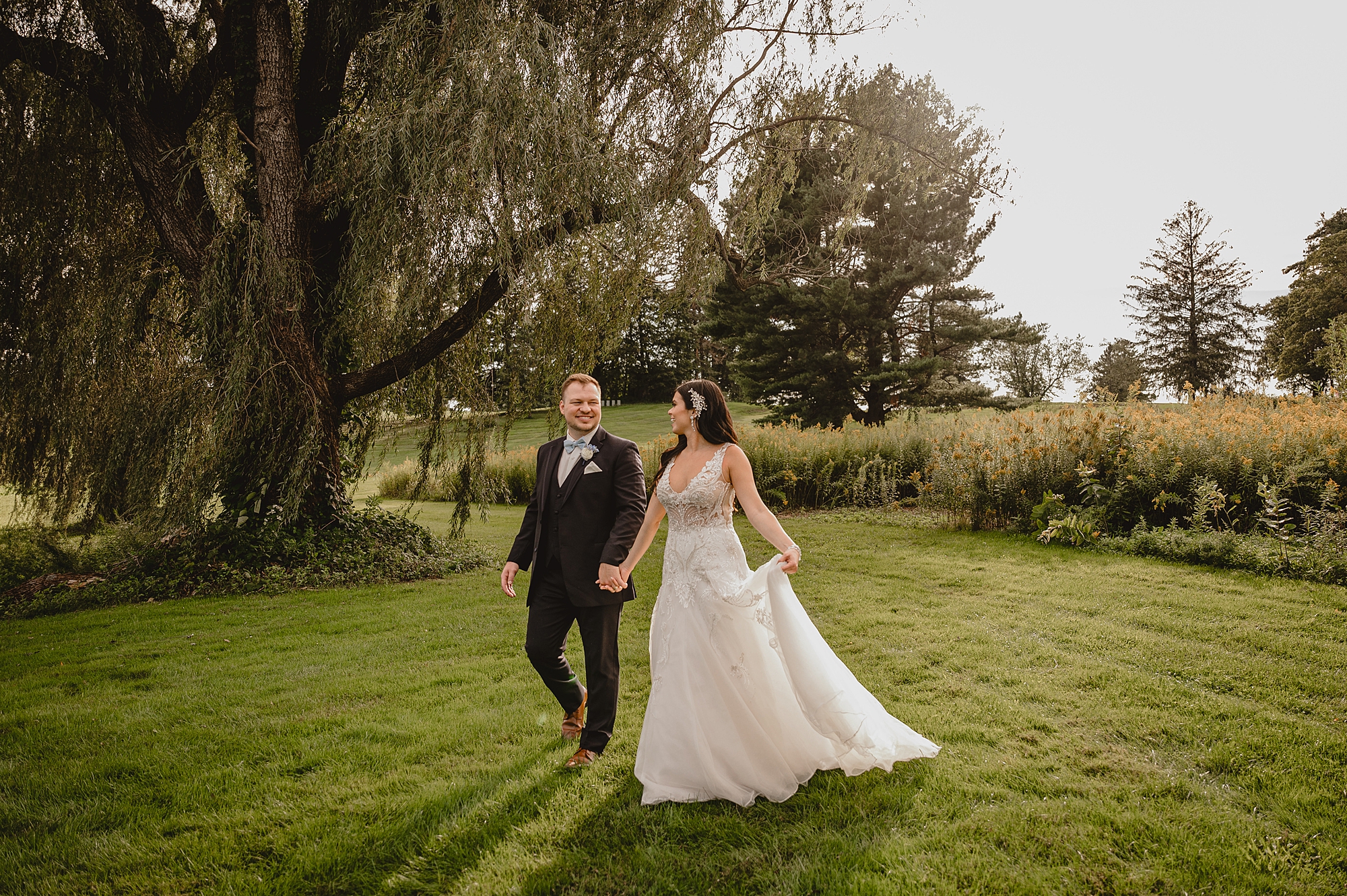 bride and groom hold hands walking by oak tree