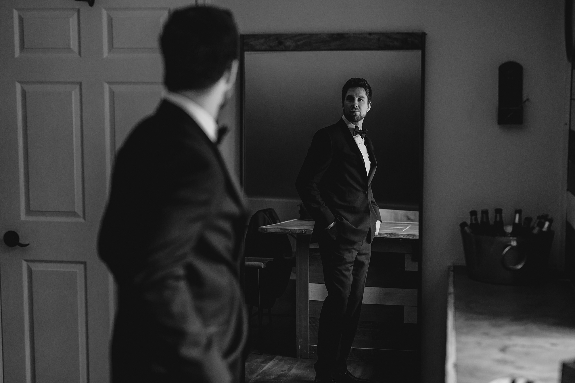 groom stands in mirror looking at reflection in black suit