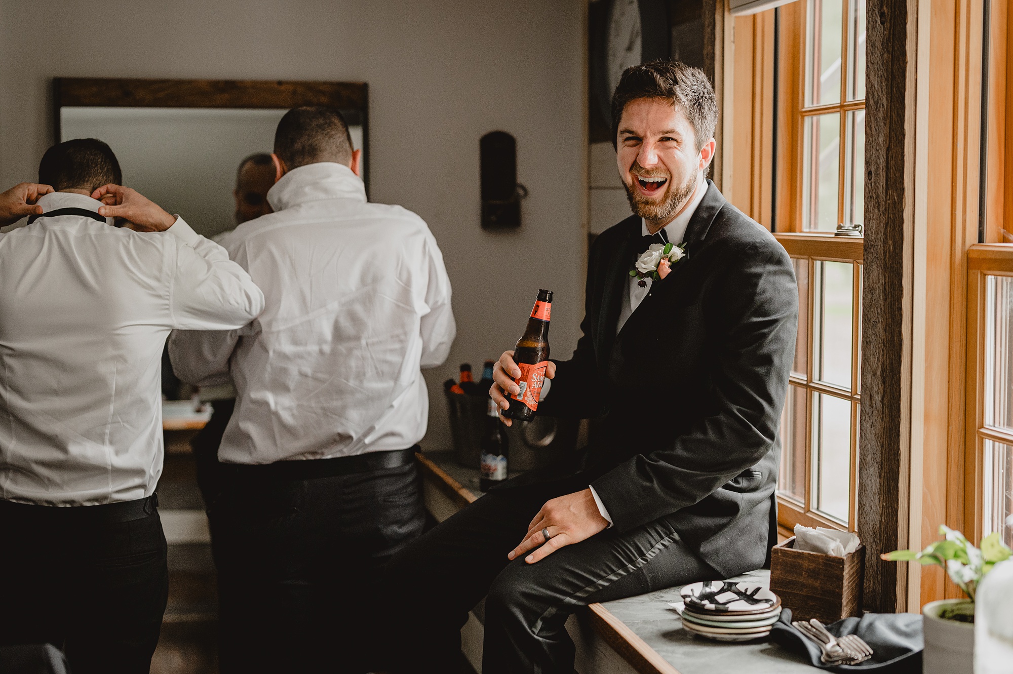 groom sits on window sill drinking beer on wedding day