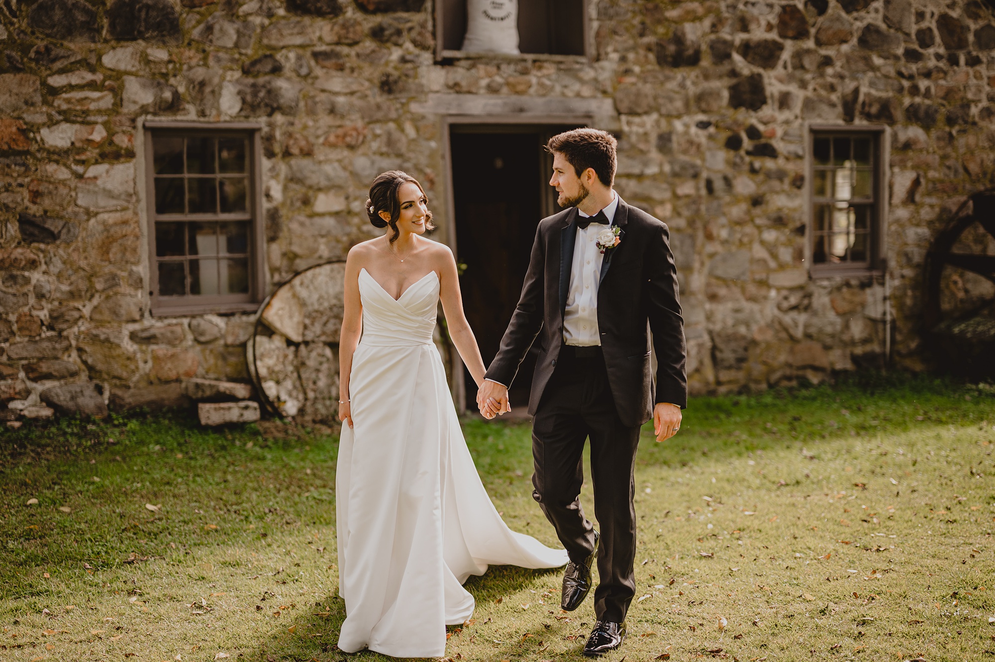 bride and groom hold hands walking away from stone barn in New Jersey 
