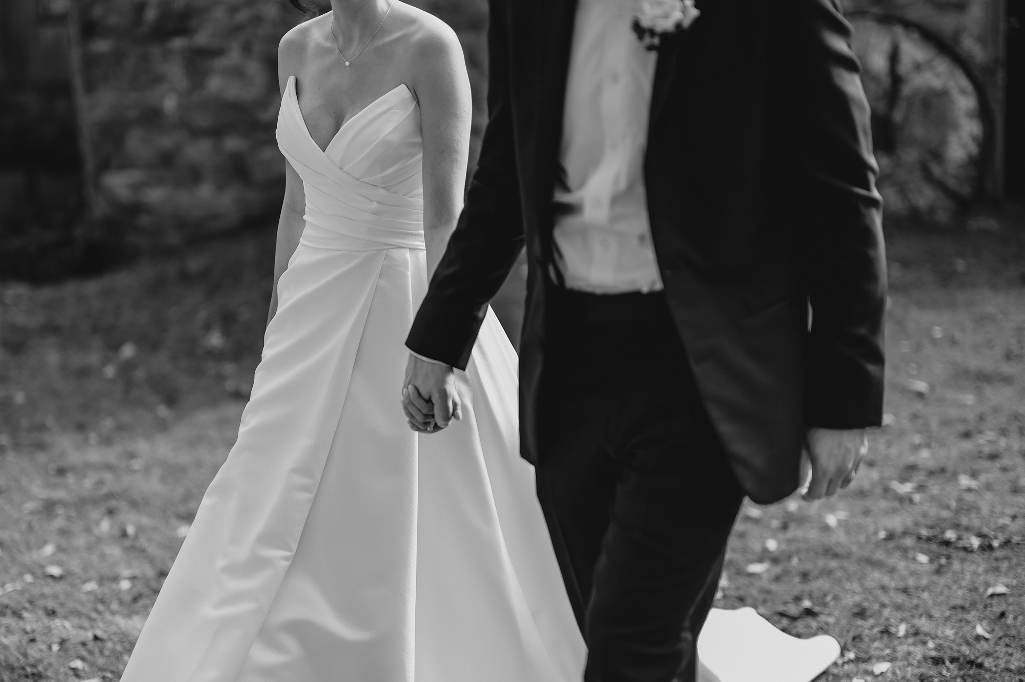groom in black tux holds bride's hand walking on lawn 