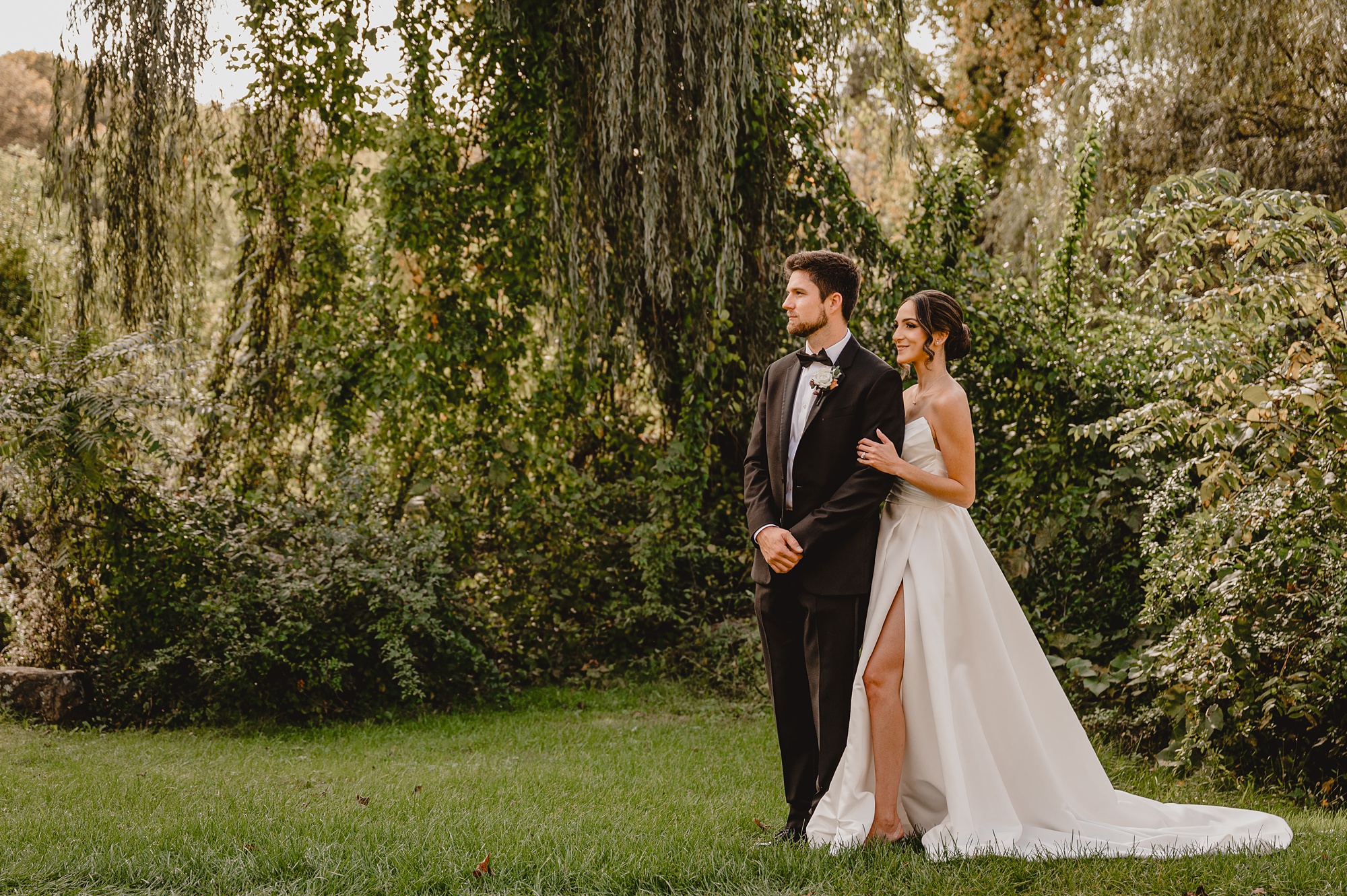 bride stands behind groom holding his arm under willow tree 