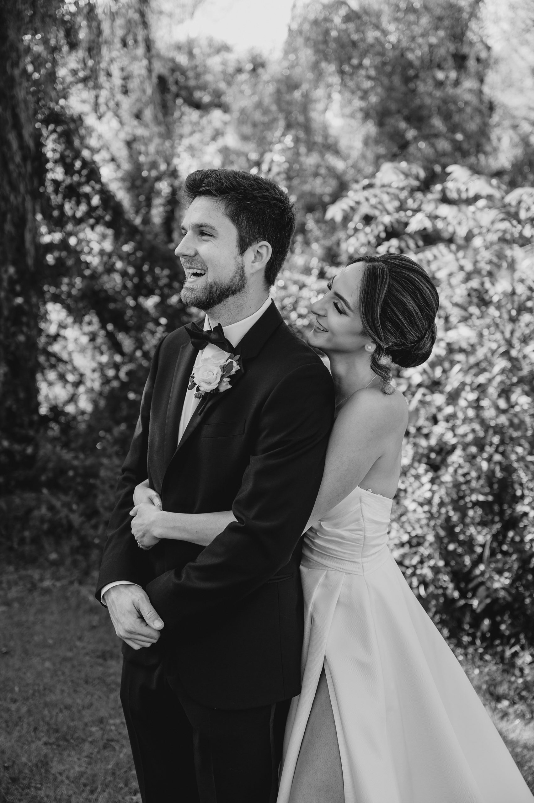 bride hugs groom around waist smiling at him under weeping willow tree