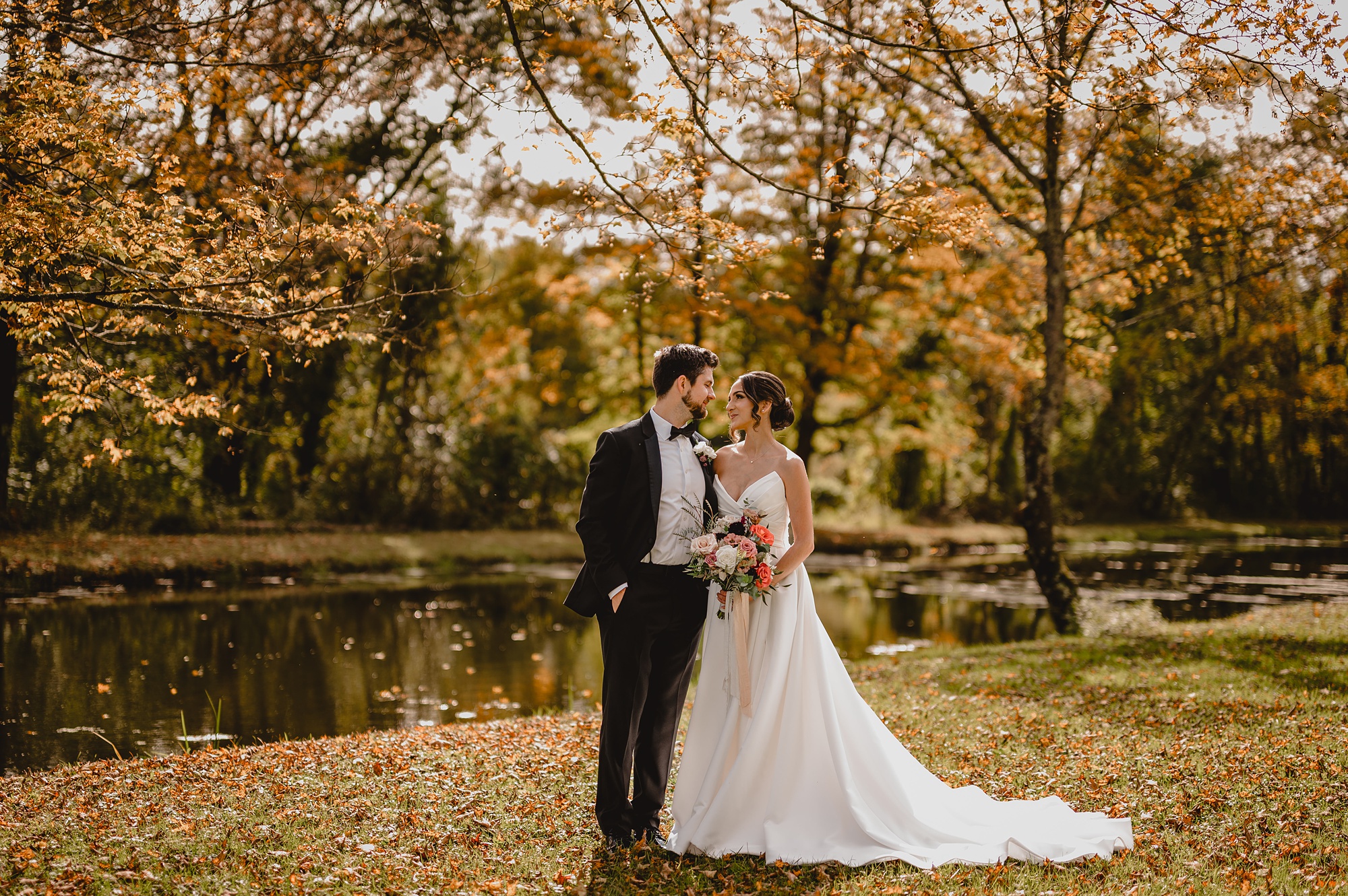 bride and groom stand looking at each other with pond behind them 