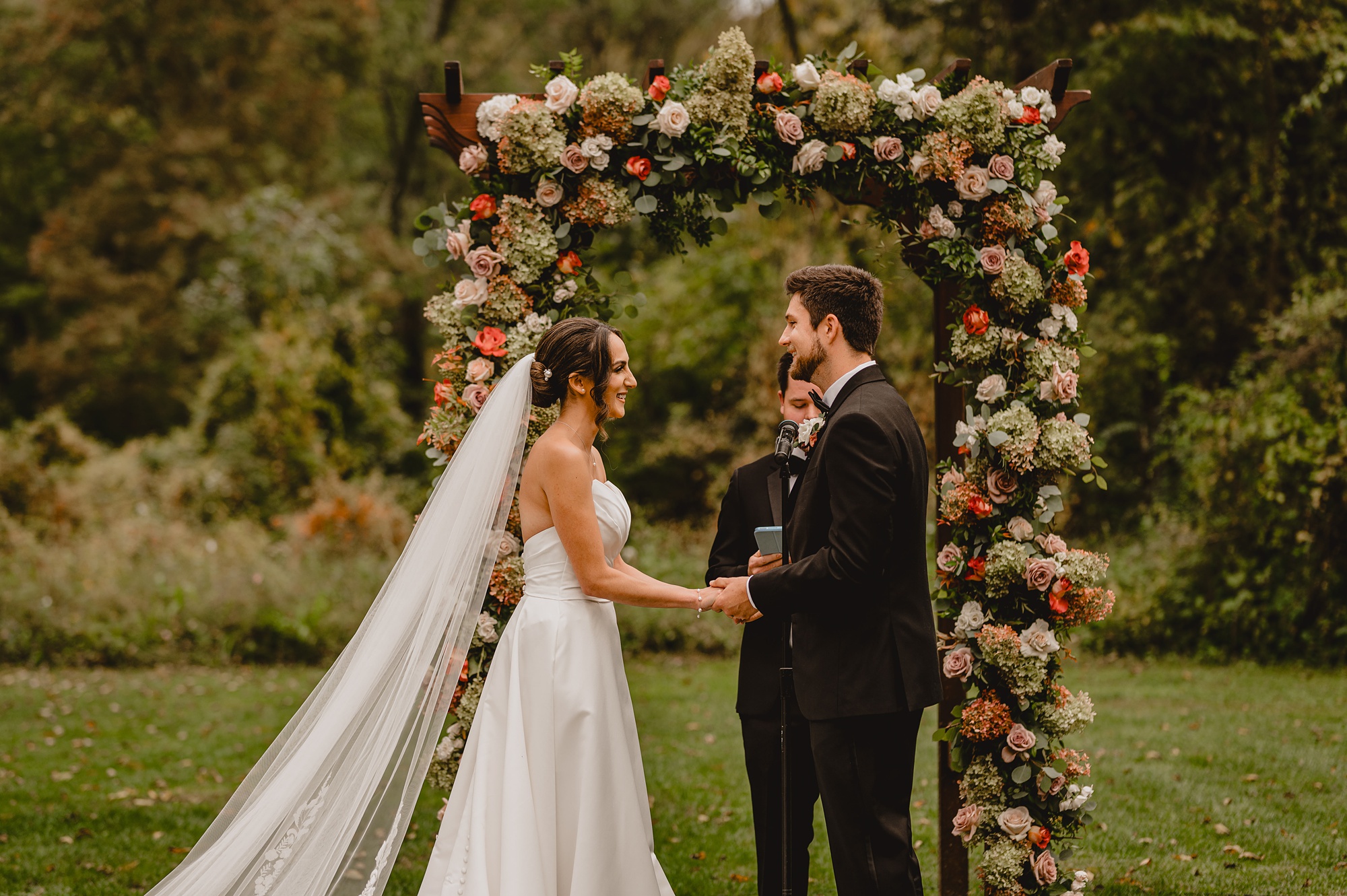 bride and groom hold hands by floral arbor during ceremony at Waterloo Village