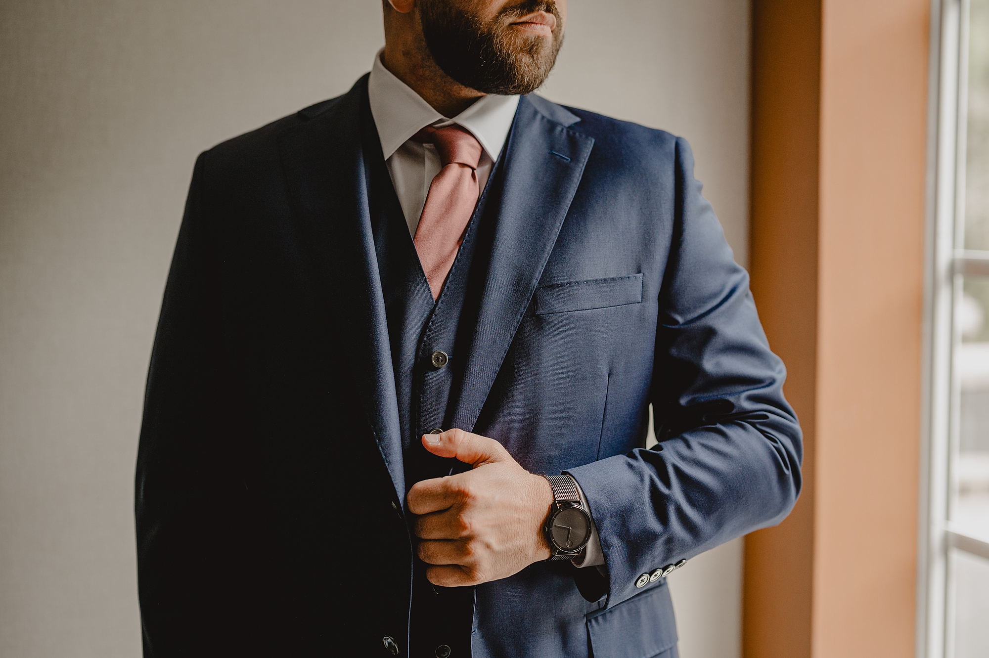 groom holds lapel of navy suit with orange tie