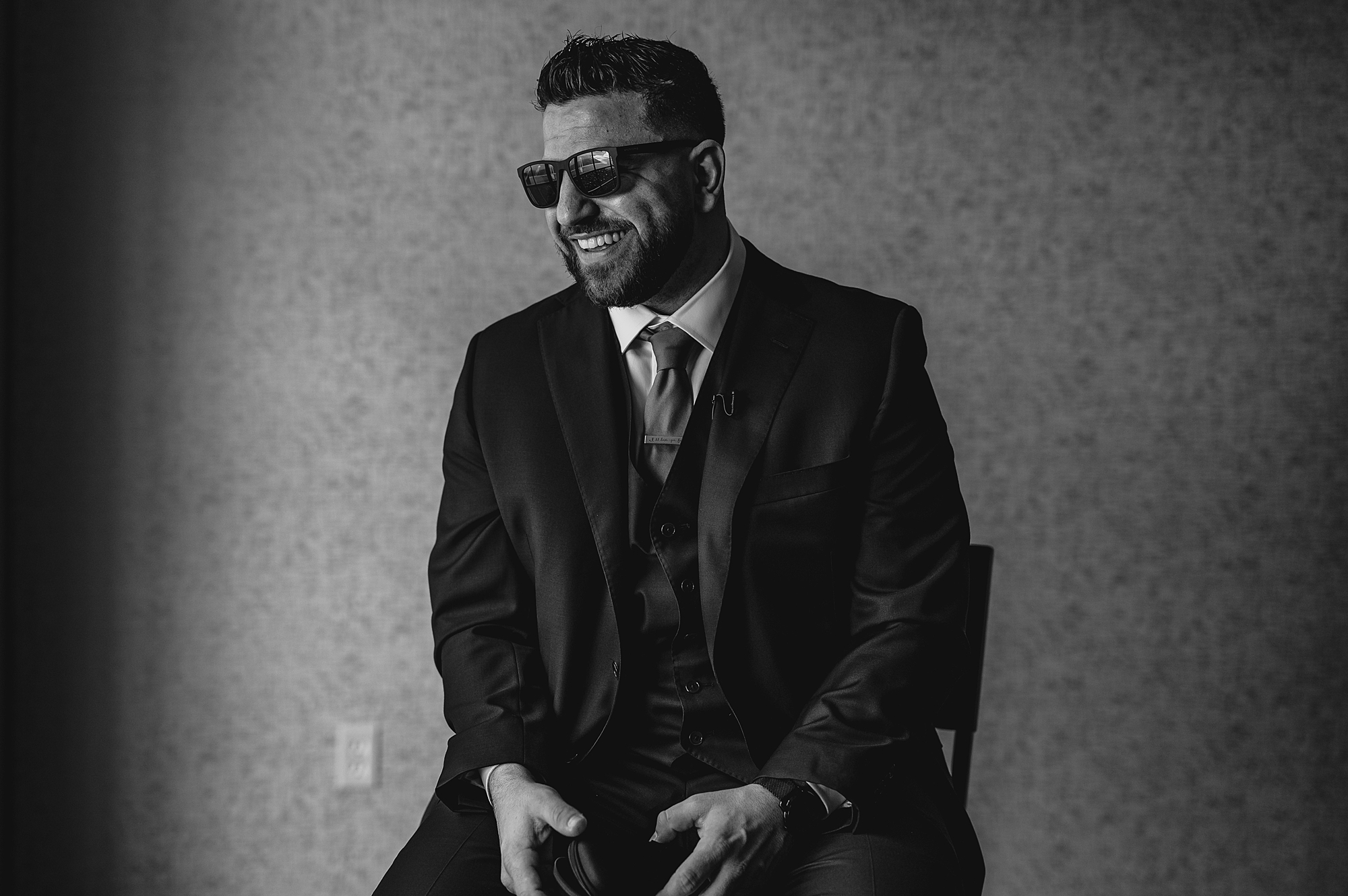 groom sits laughing in suit with sunglasses