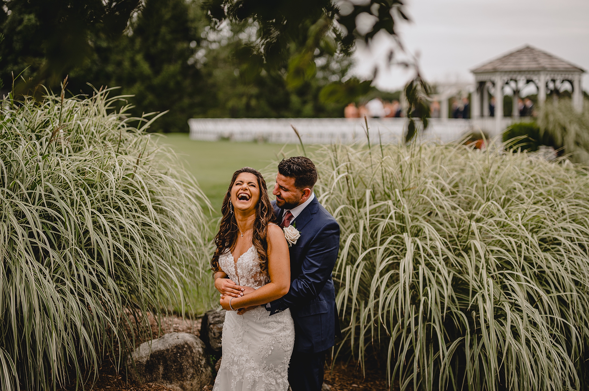 groom hugs bride from behind making her laugh between big green bushes