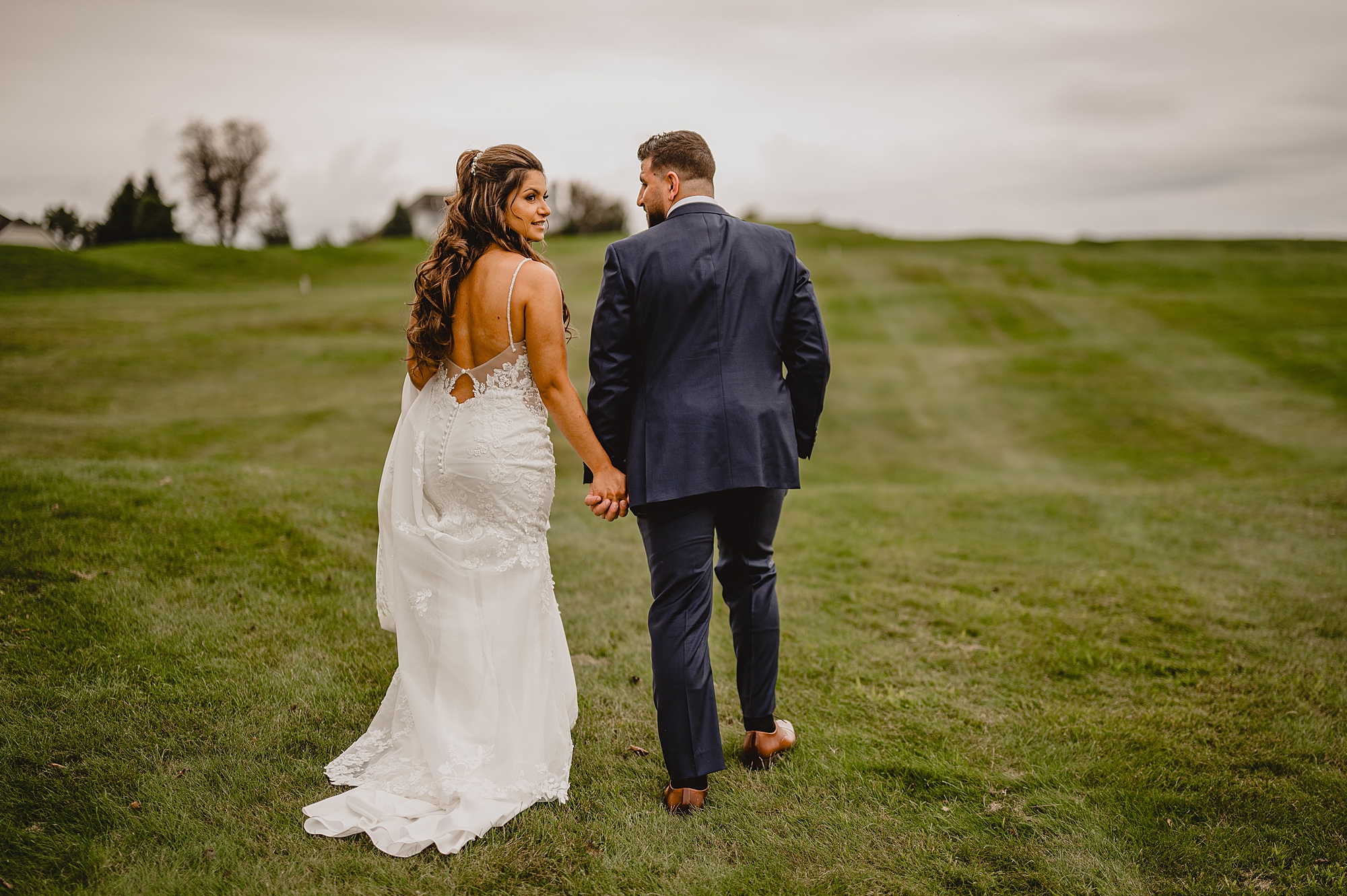 bride and groom hold hands walking on hill while bride looks over shoulder