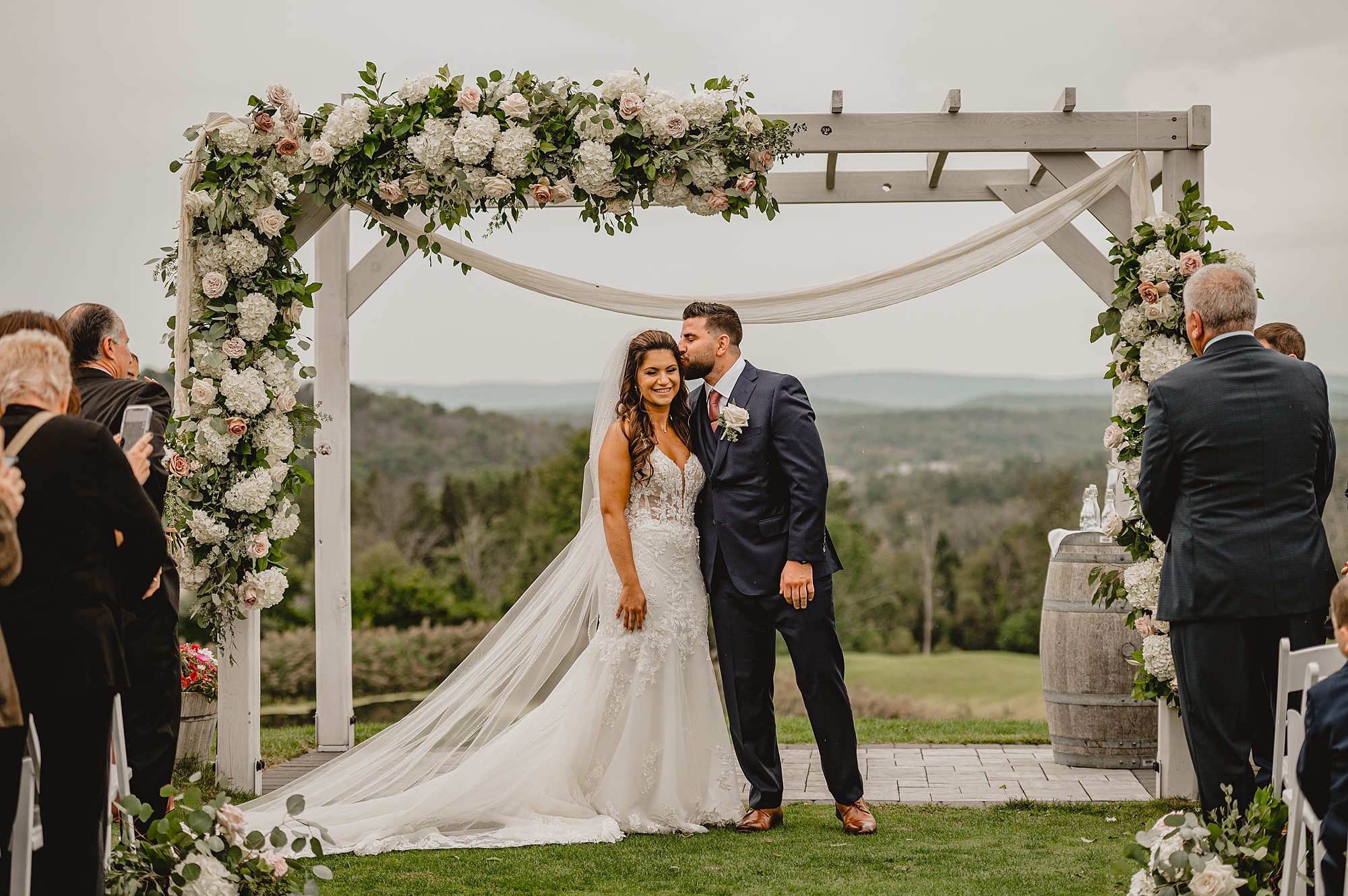 groom kisses bride's forehead under wooden arbor with flowers during ceremony on lawn at Skyview Golf Club