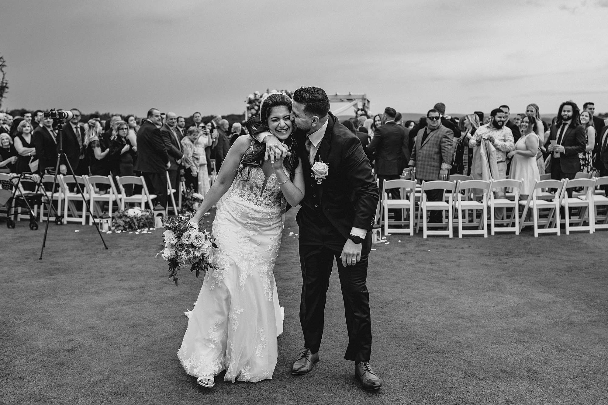 groom hugs bride around the neck kissing her cheek leaving ceremony on lawn at Skyview Golf Club