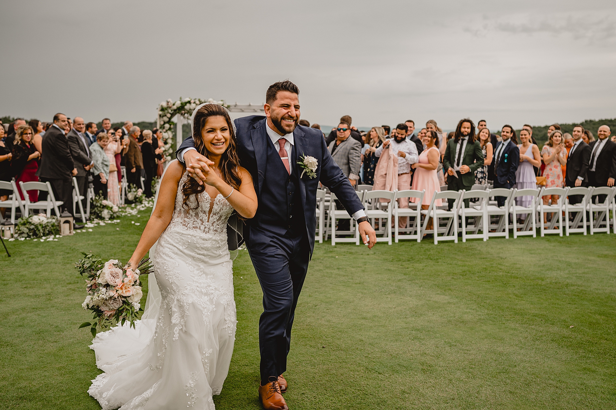 bride and groom walk away from Ariel after ceremony on lawn at Skyview Golf Club