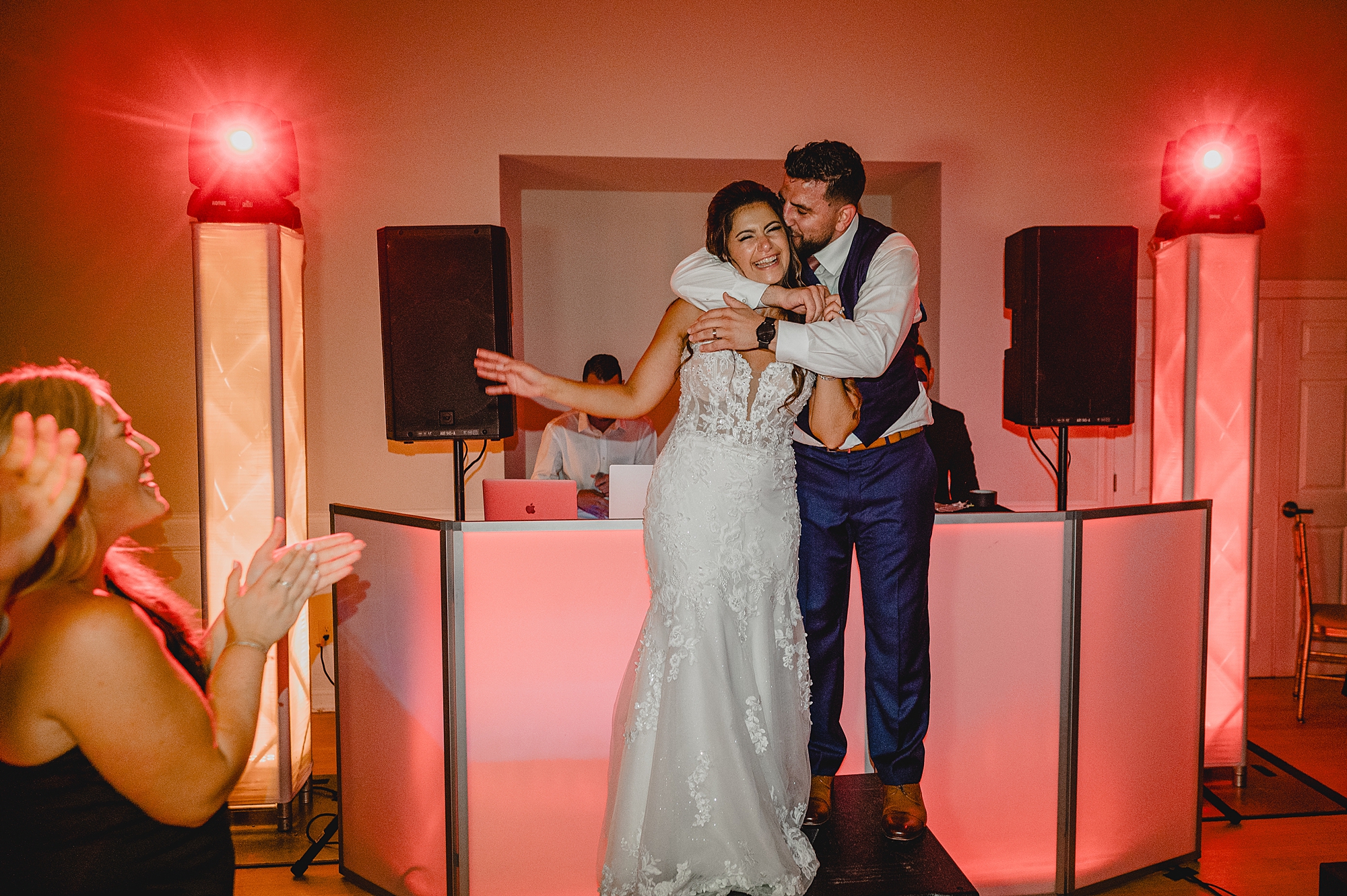groom hugs bride from behind in front of DJ stand for New Jersey wedding reception