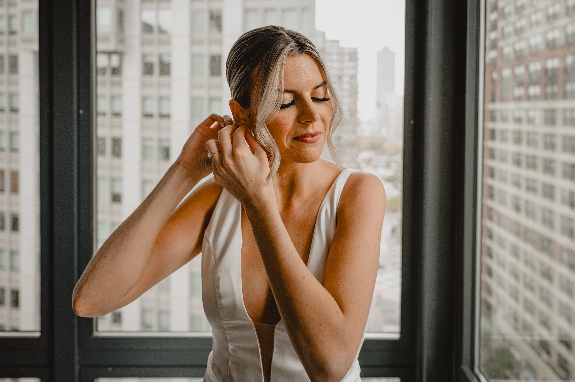 blonde woman adjusts earrings in hotel room looking over Jersey City