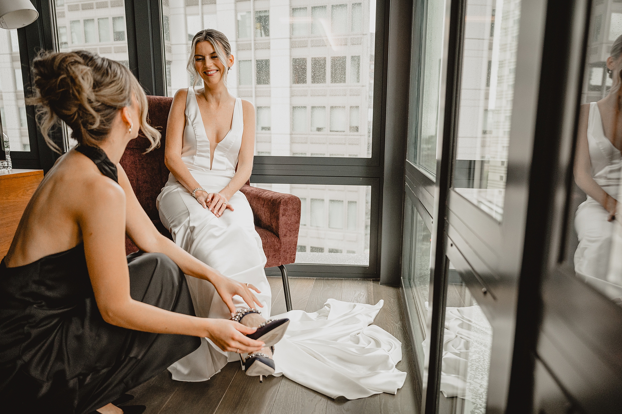 bridesmaid helps bride with shoes in front of windows of Jersey City hotel room