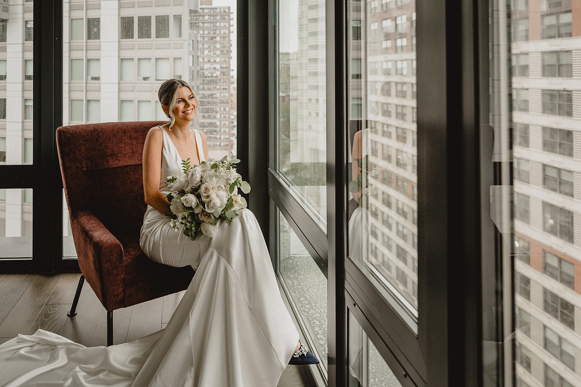 bride sits in velvet chair in front of wall of windows in Jersey City hotel room