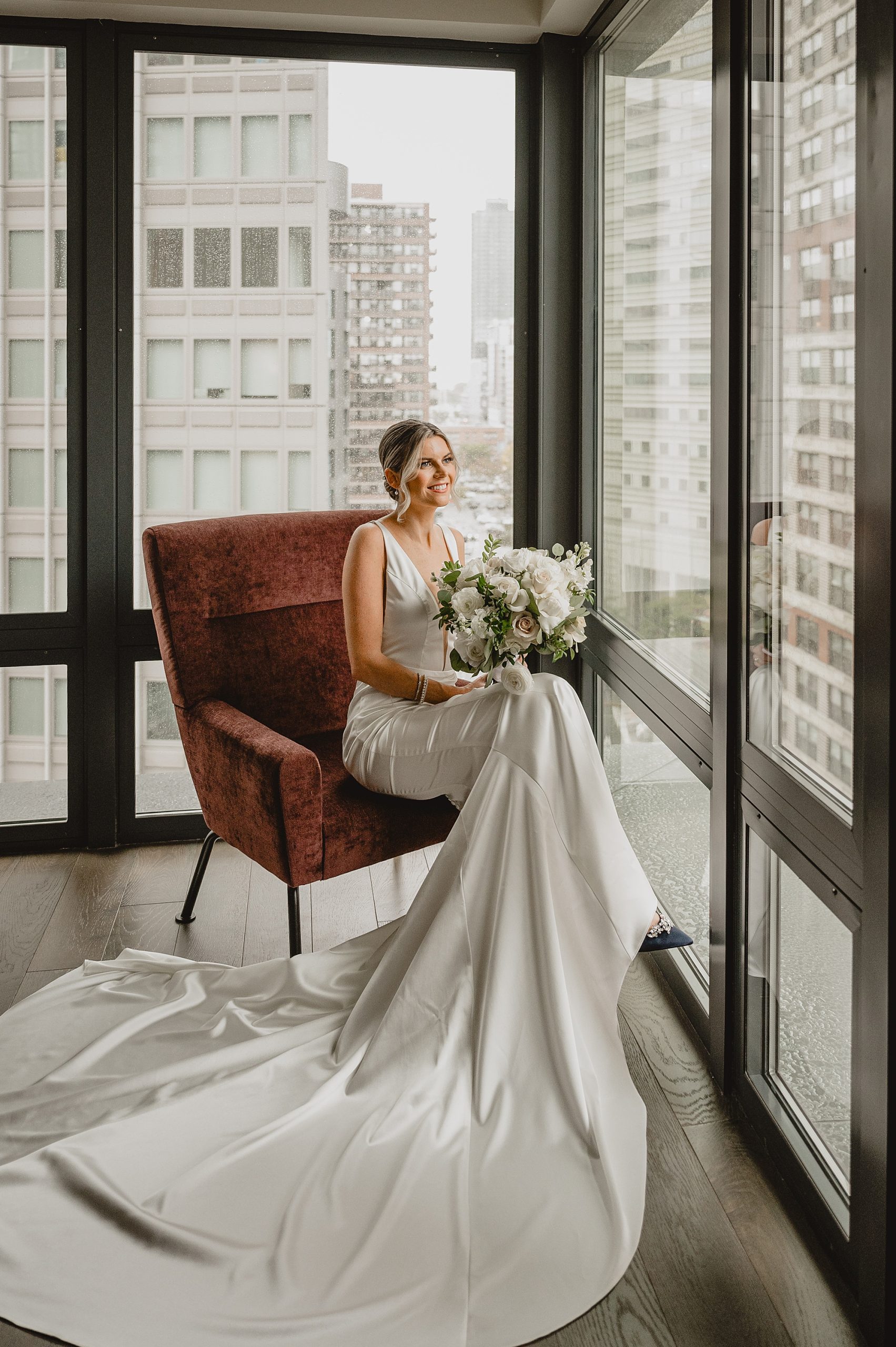 bride sits in velvet chair in front of wall of windows in Jersey City hotel room