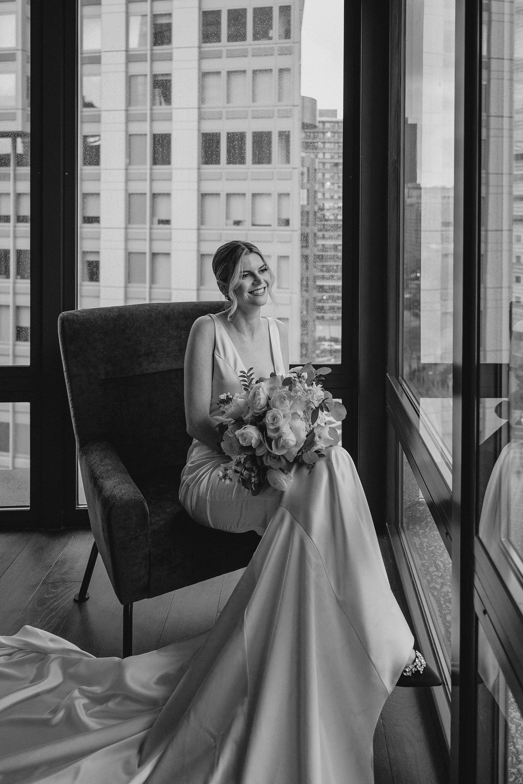 bride sits in velvet chair in front of wall of windows in Jersey City hotel room