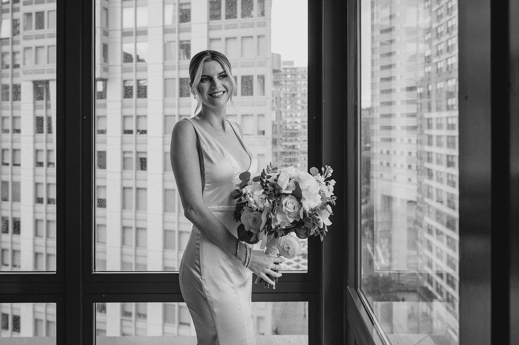 bride stands holding bouquet of white flowers in front of wall of windows