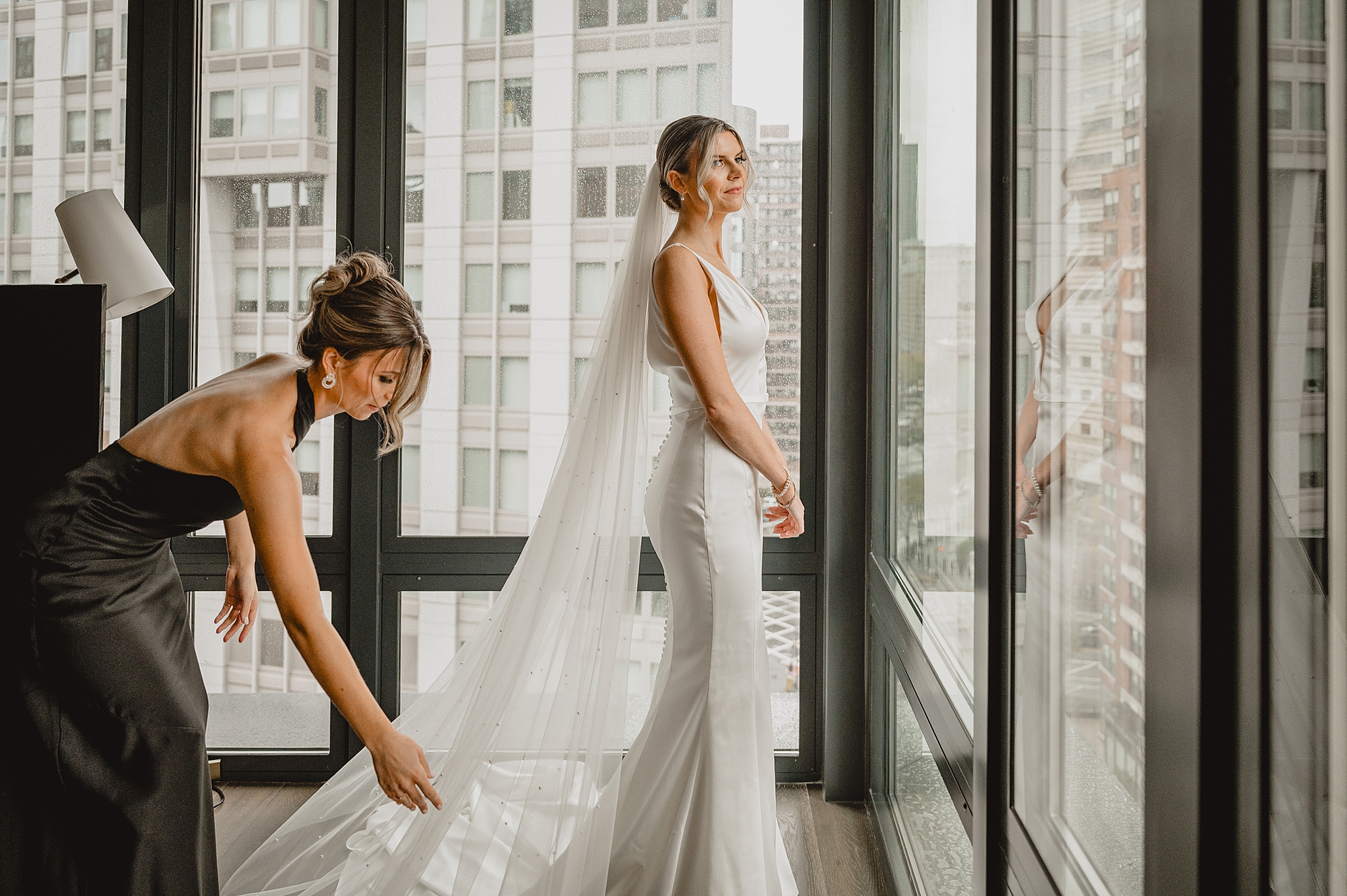 bridesmaid helps bride with veil behind her in hotel room in Jersey City
