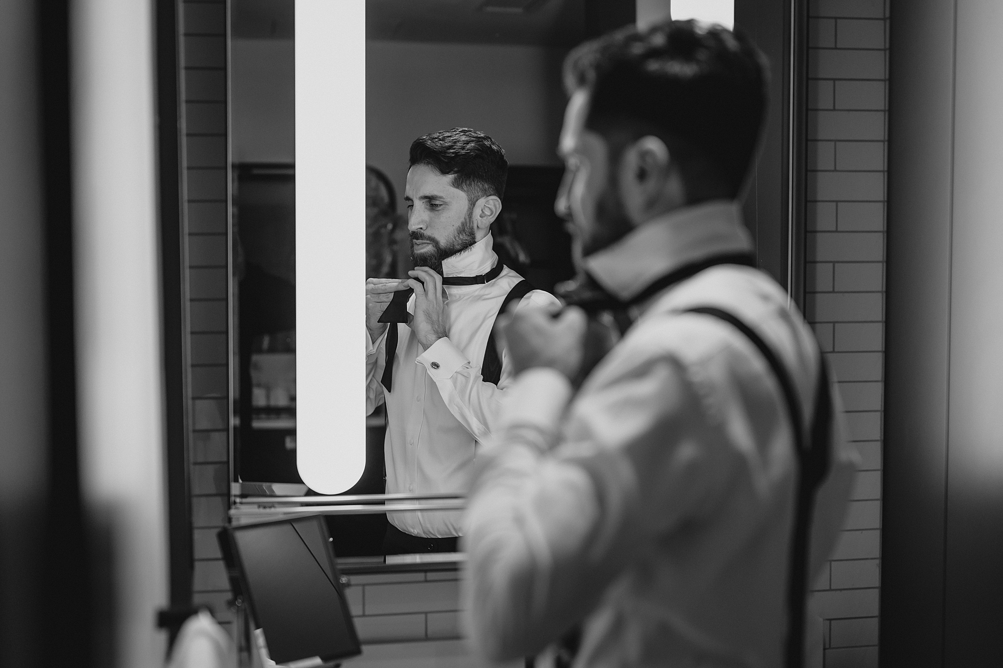 groom adjusts black bowtie in mirror of Jersey City hotel