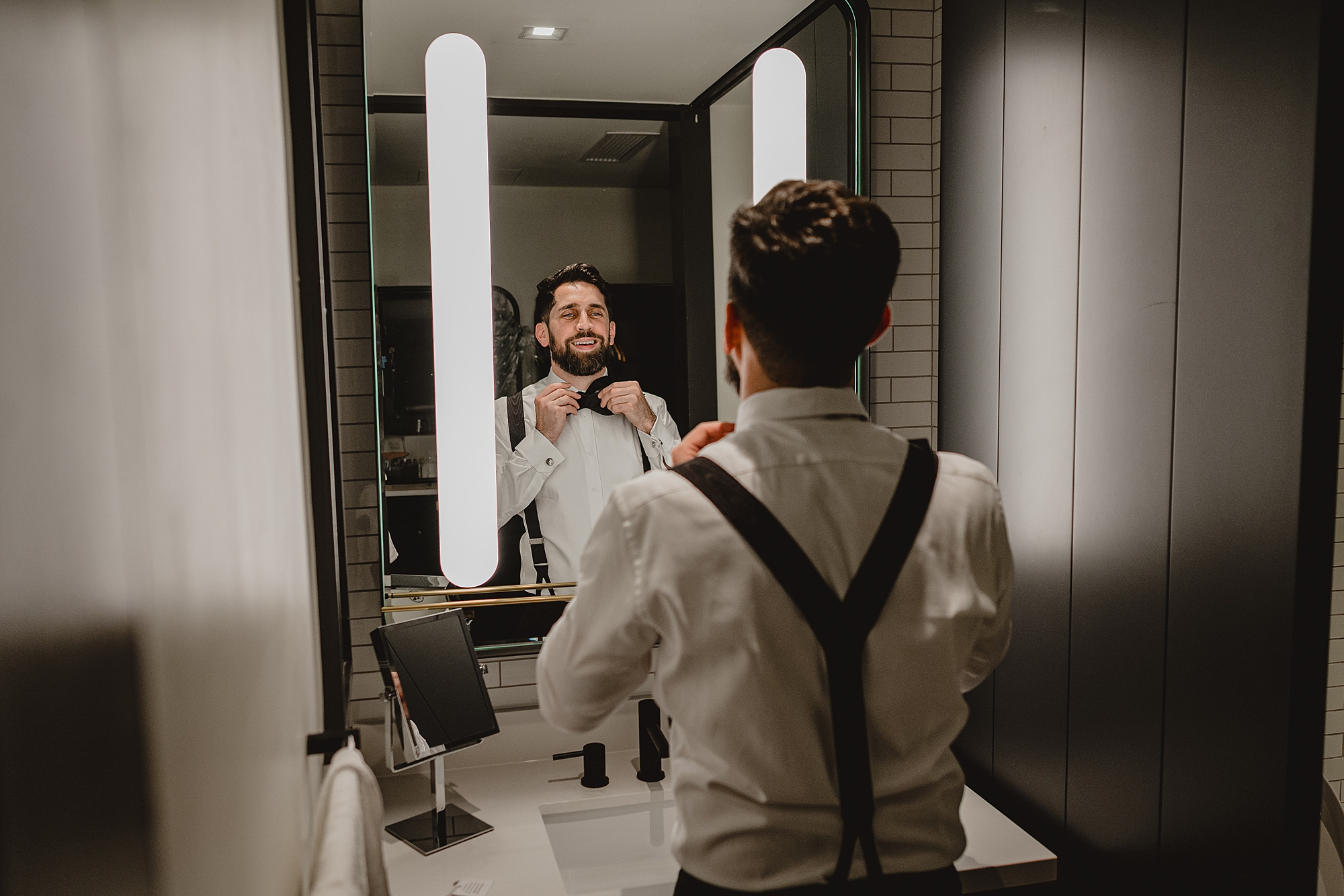 groom adjusts bowtie in mirror of Jersey City hotel room