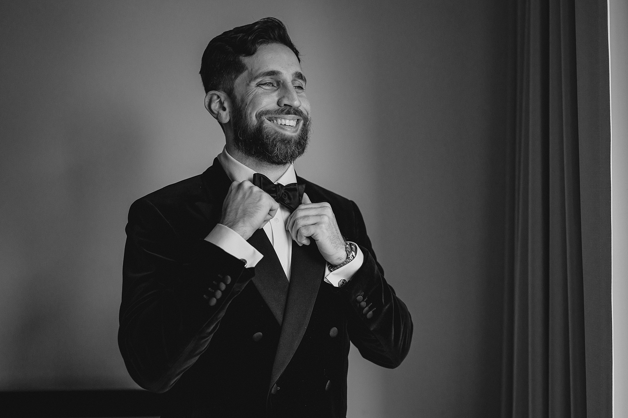 man adjusts bowtie of velvet jacket in hotel room in Jersey City