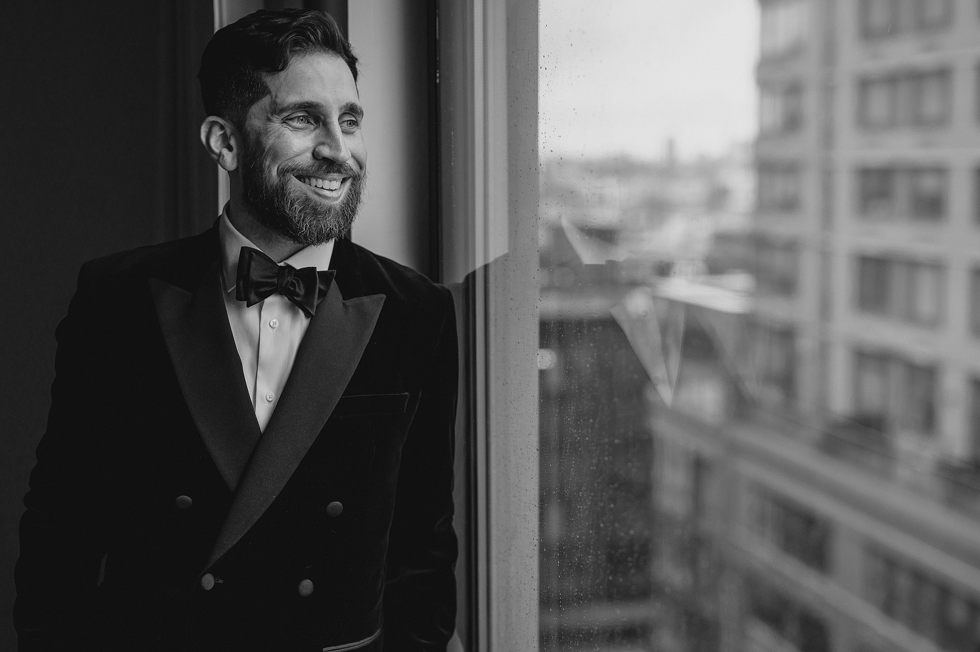 groom leans against window overlooking Jersey City in hotel room