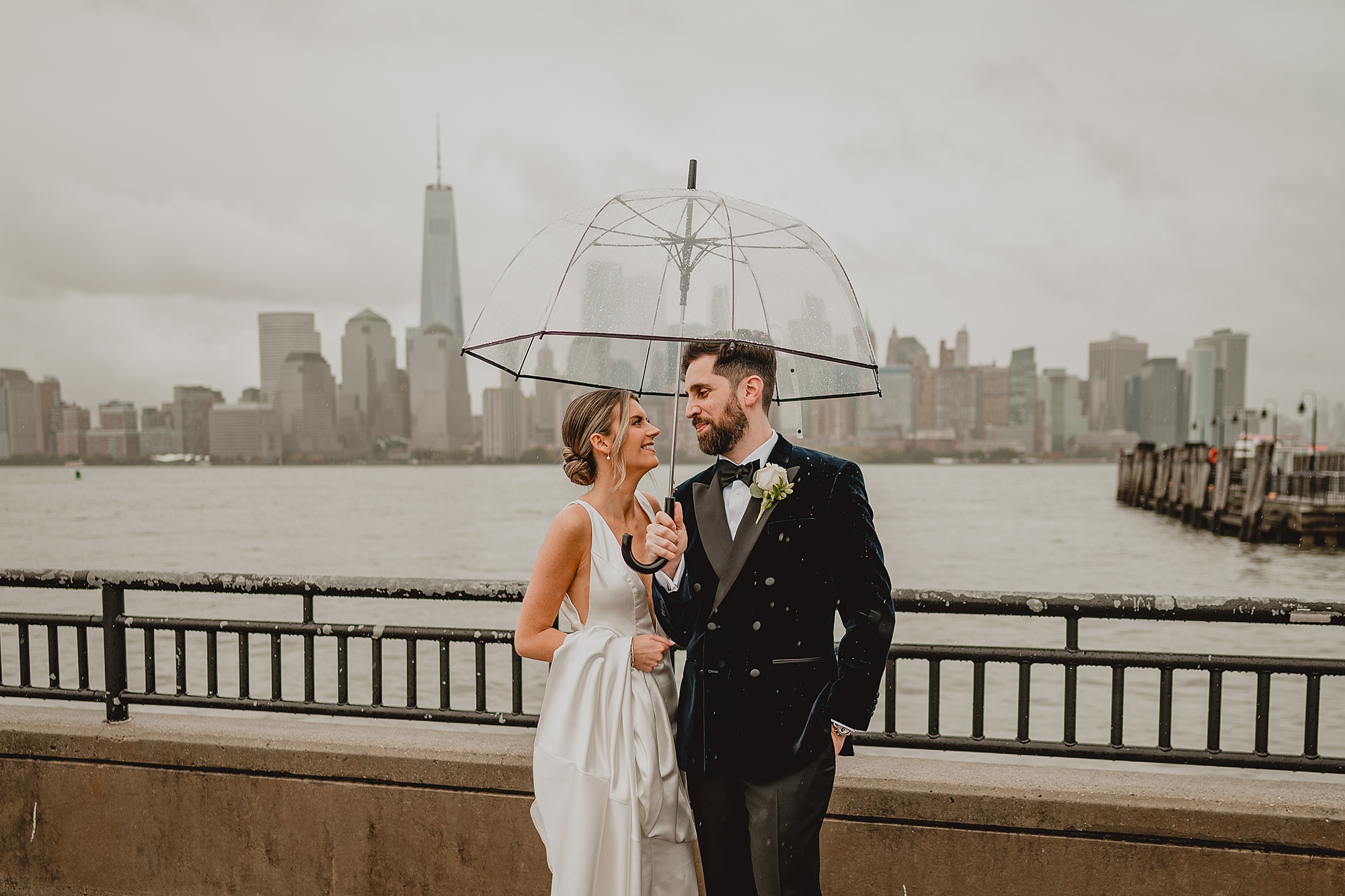 bride and groom hug under clear umbrella in front of Hudson River on rainy wedding day