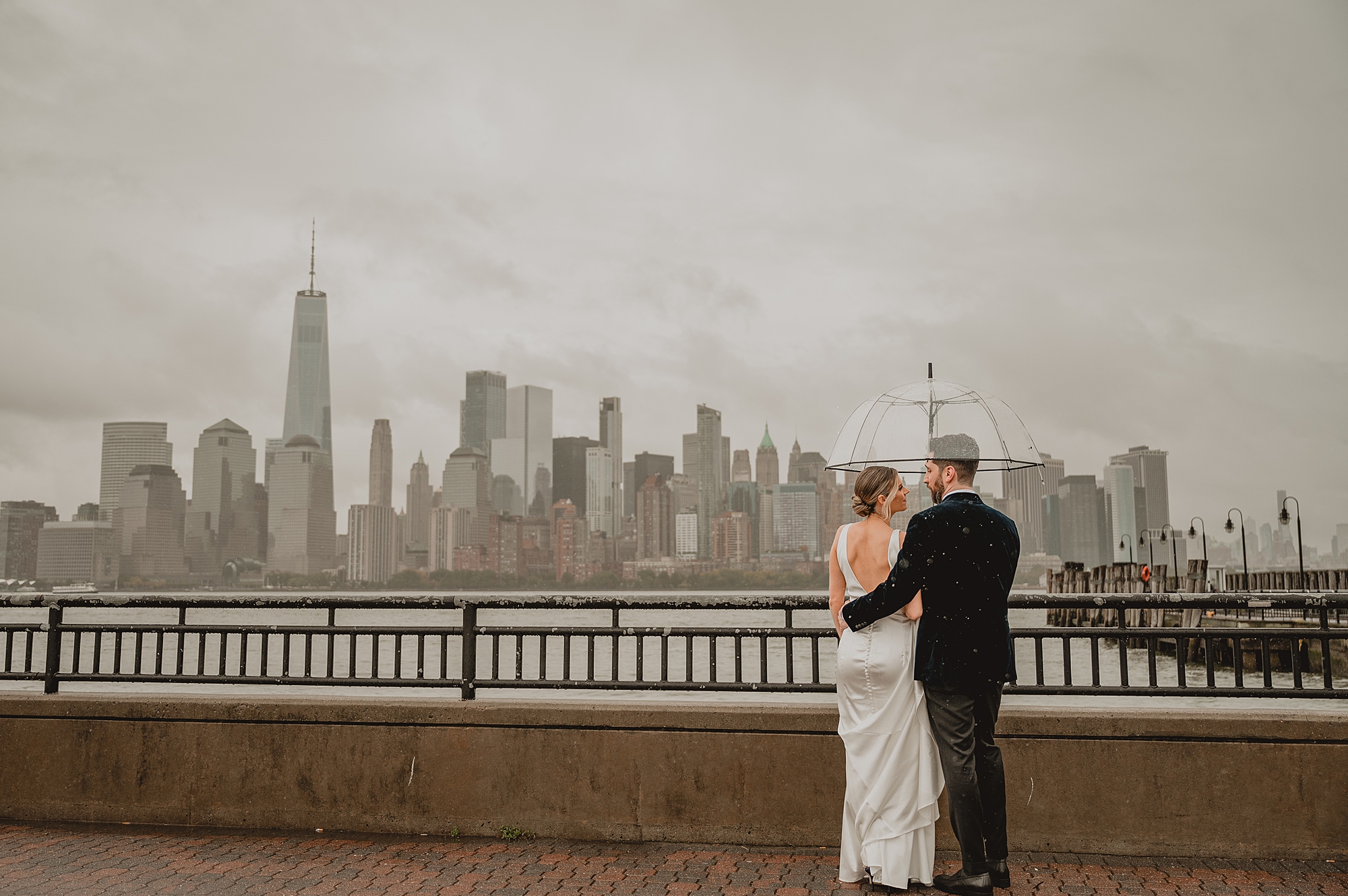 newlyweds hug under clear umbrella in front of Hudson River outside Maritime Parc