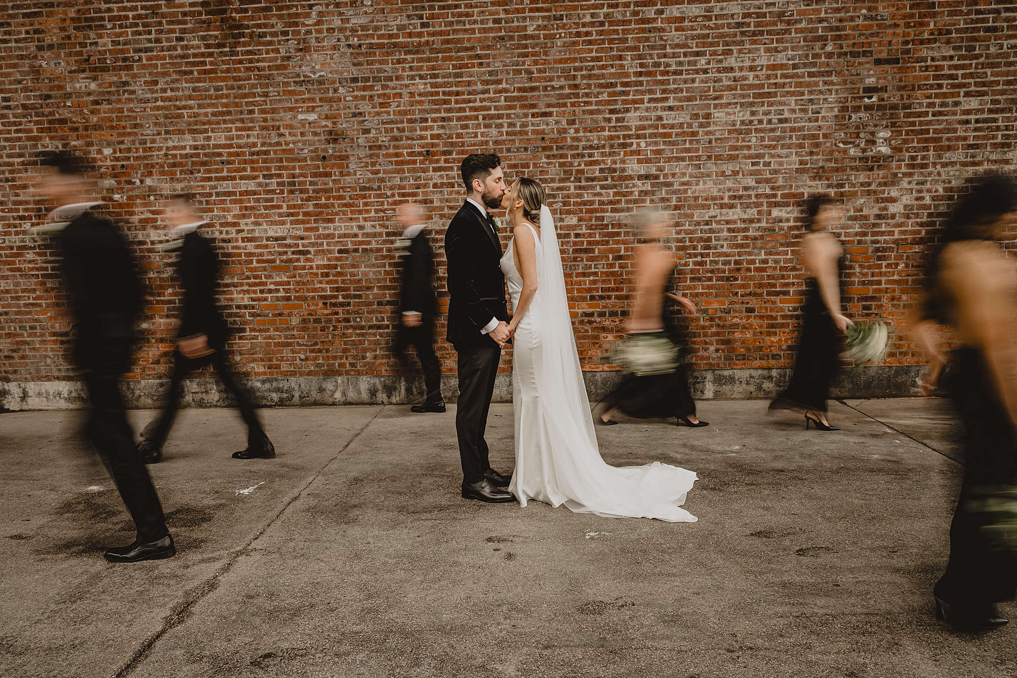 bride and groom kiss while bridal party walks around them in front of brick wall