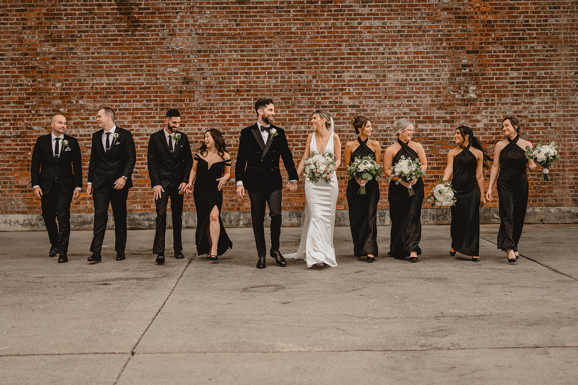 newlyweds walk with bridesmaids and groomsmen in black attire in front of brick wall at Maritime Parc