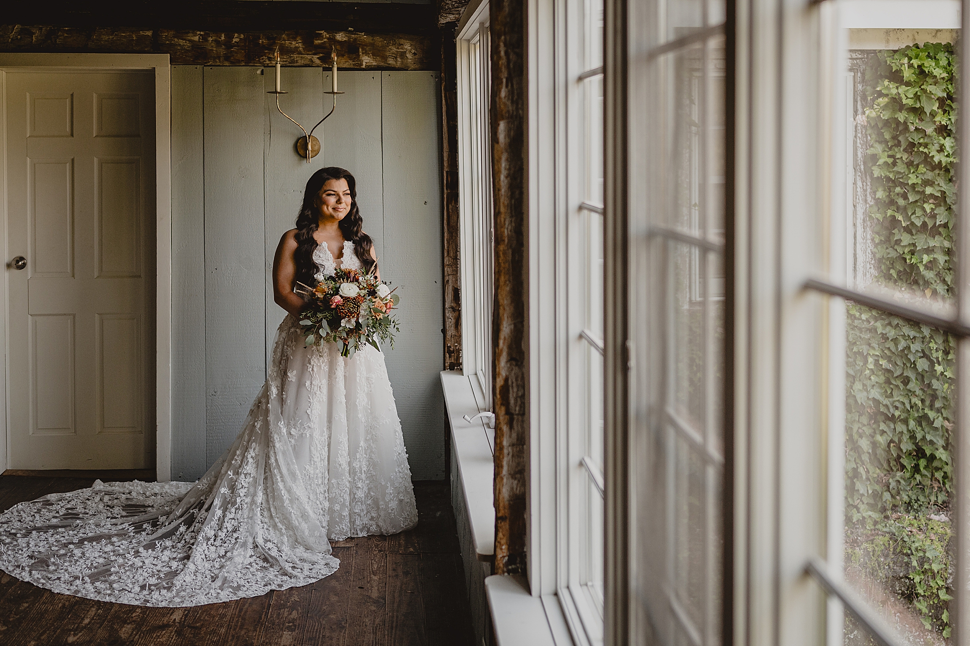 bride stands holding fall bouquet looking out windows
