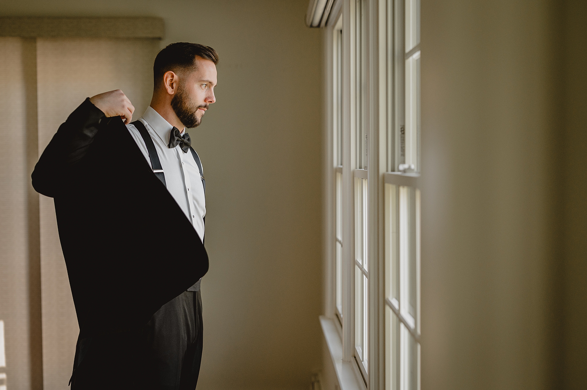 groom puts on black jacket by windows at Crossed Keys Estate