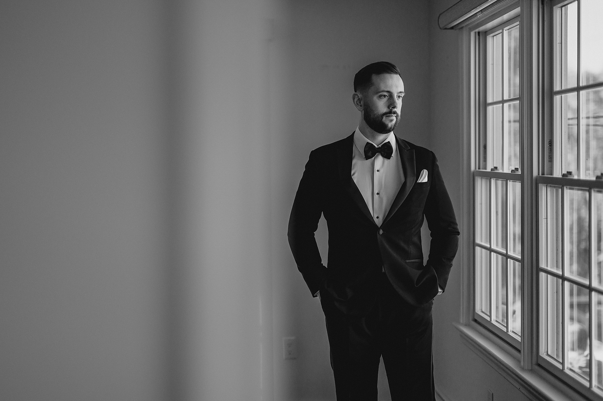 groom stands with hands in pockets looking out windows at Crossed Keys Estate