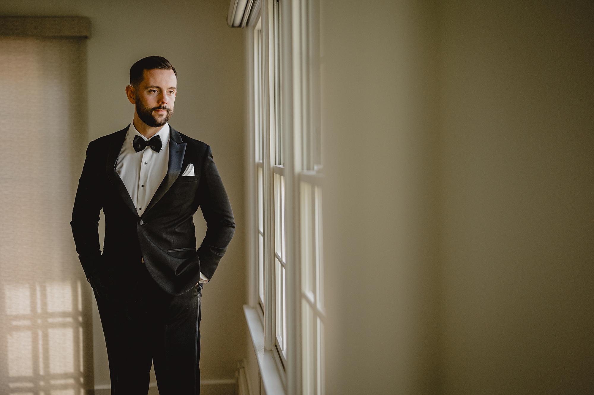 groom stands with hands in pockets looking out windows at Crossed Keys Estate