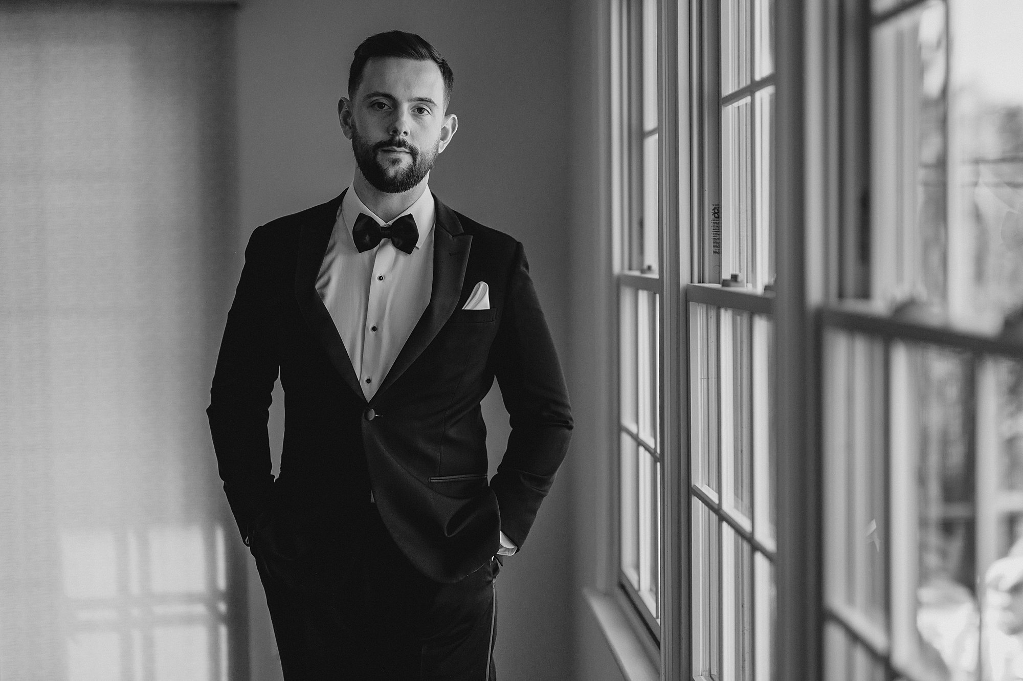 groom stands with hands in pockets looking out windows at Crossed Keys Estate