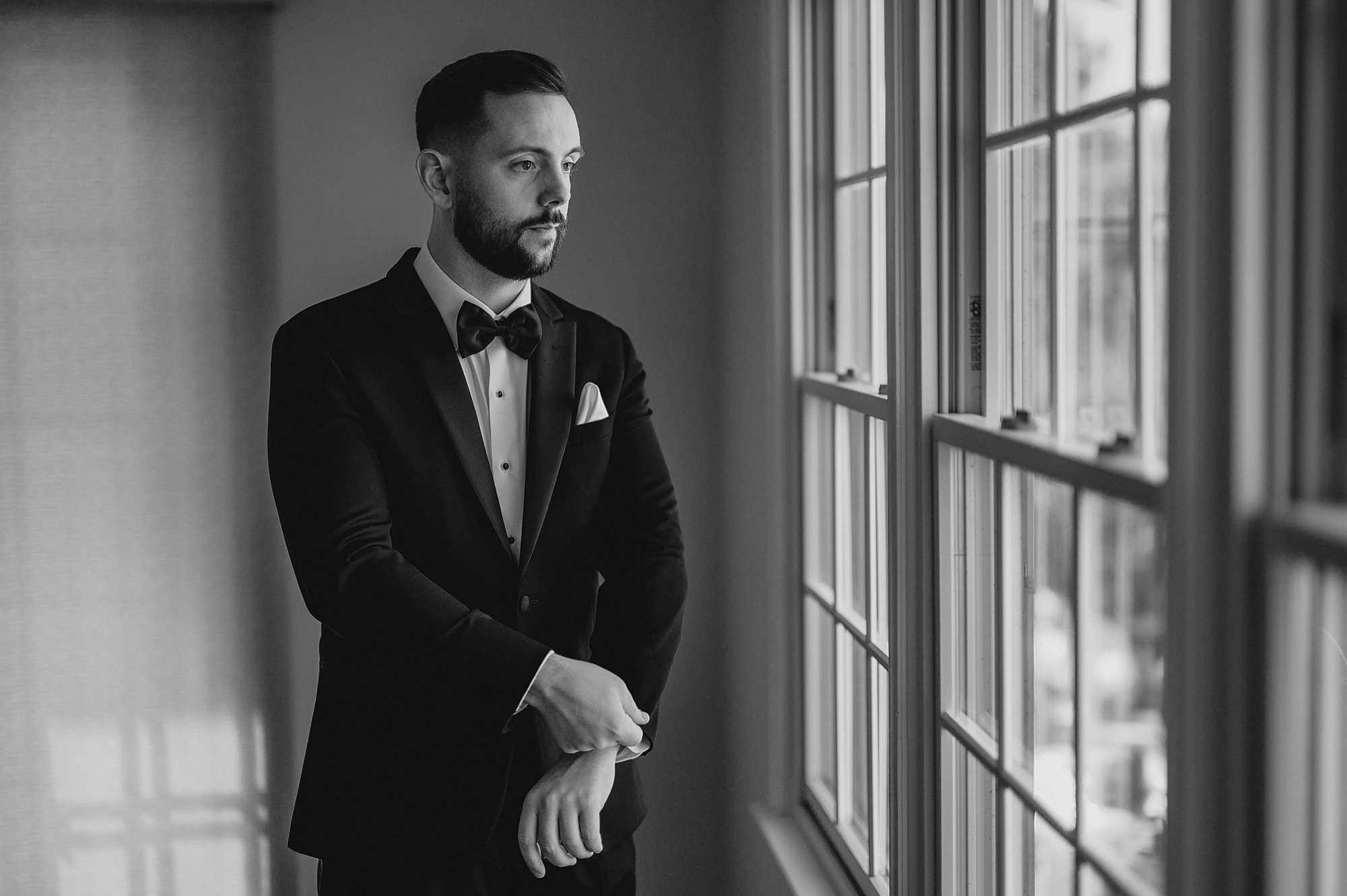 groom adjusts cufflinks of suit looking out windows at Crossed Keys Estate