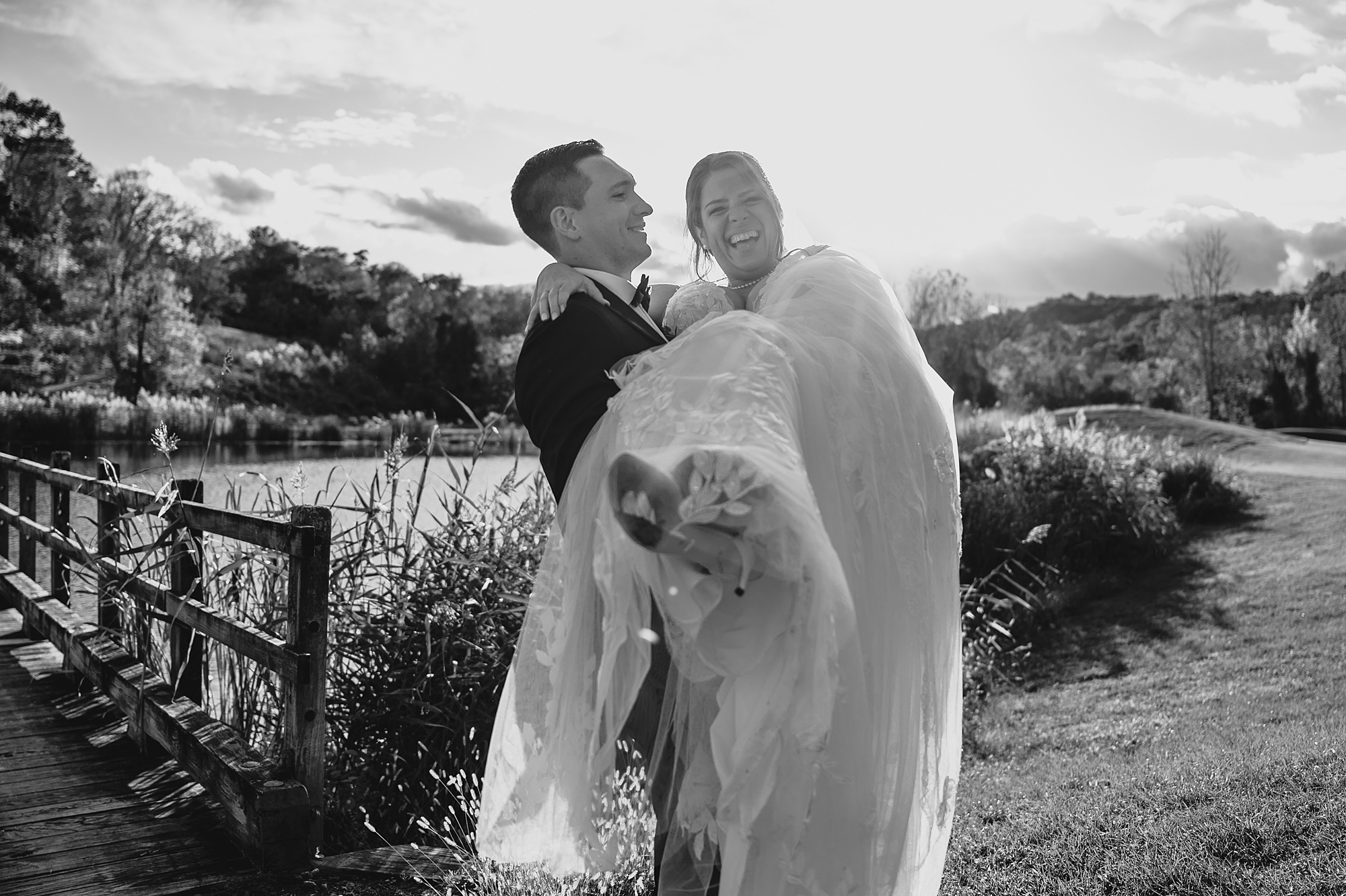 black and white photo of groom carrying bride over bridge