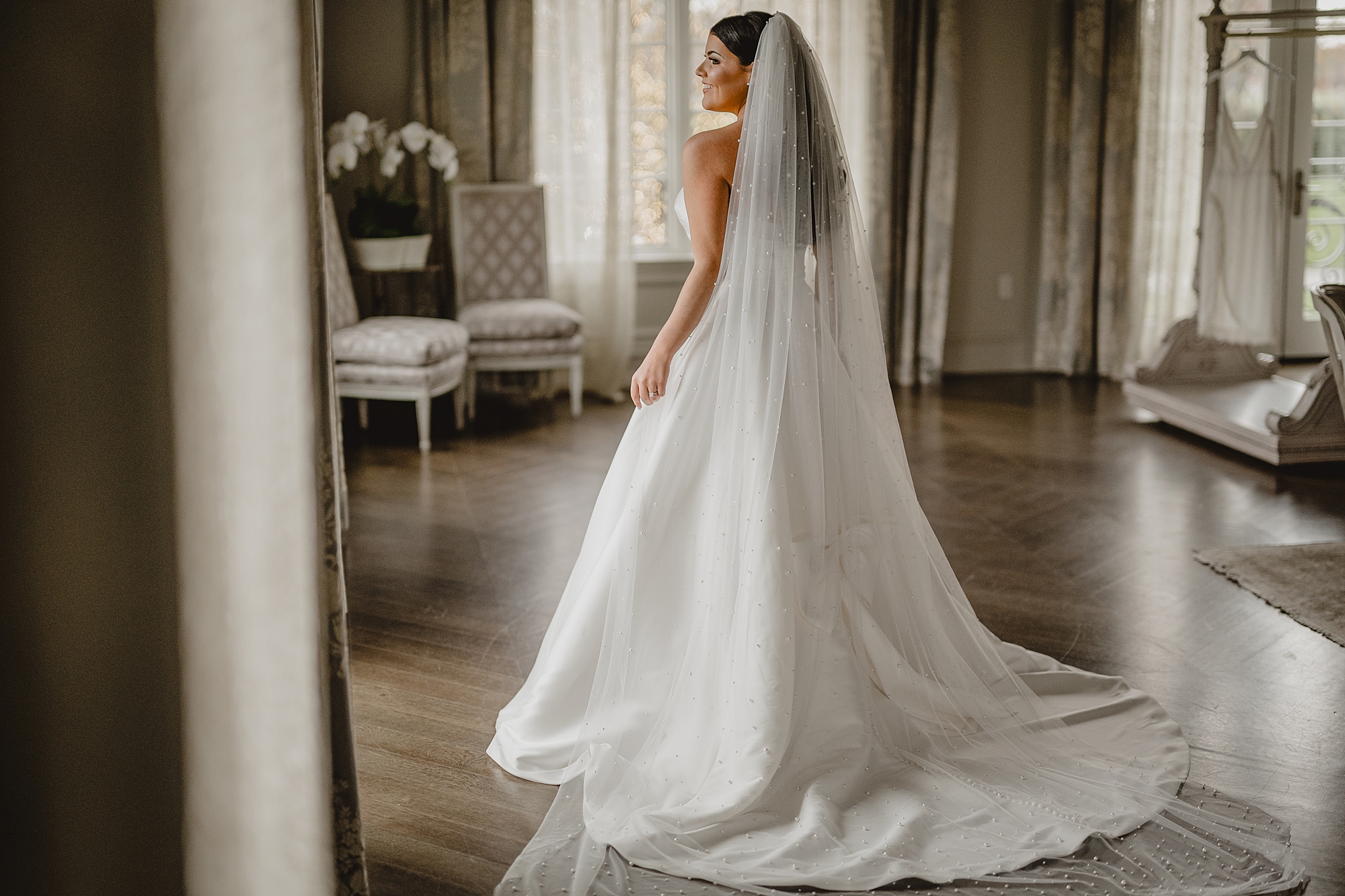 bride looks out the window with veil trailing behind her in strapless gown