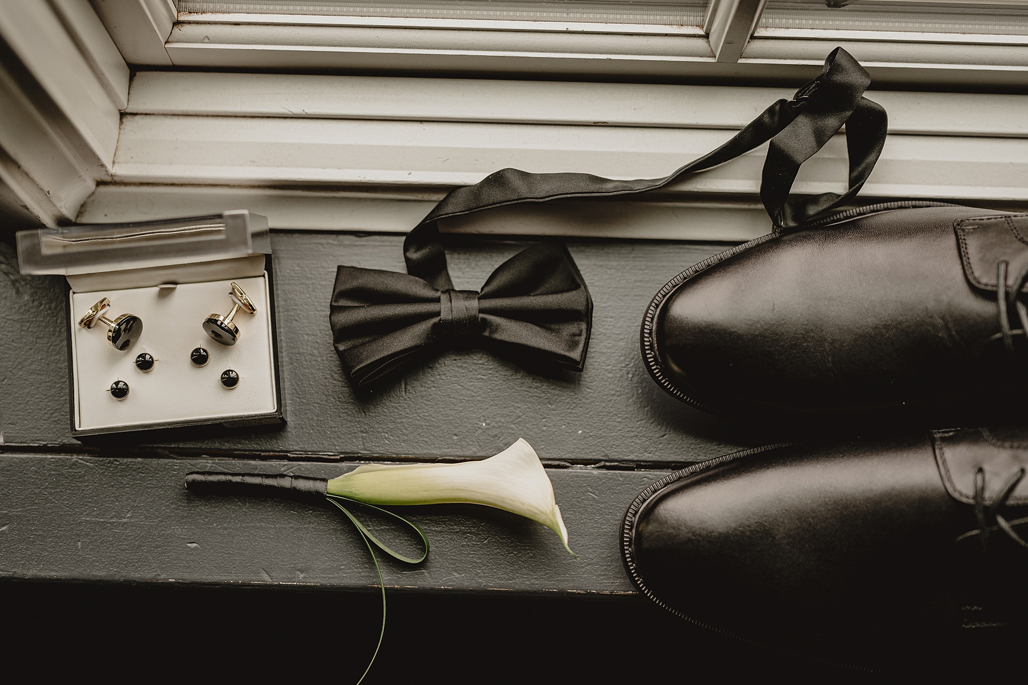 groom's black shoes and black tie sit next to calla lily on window ledge 