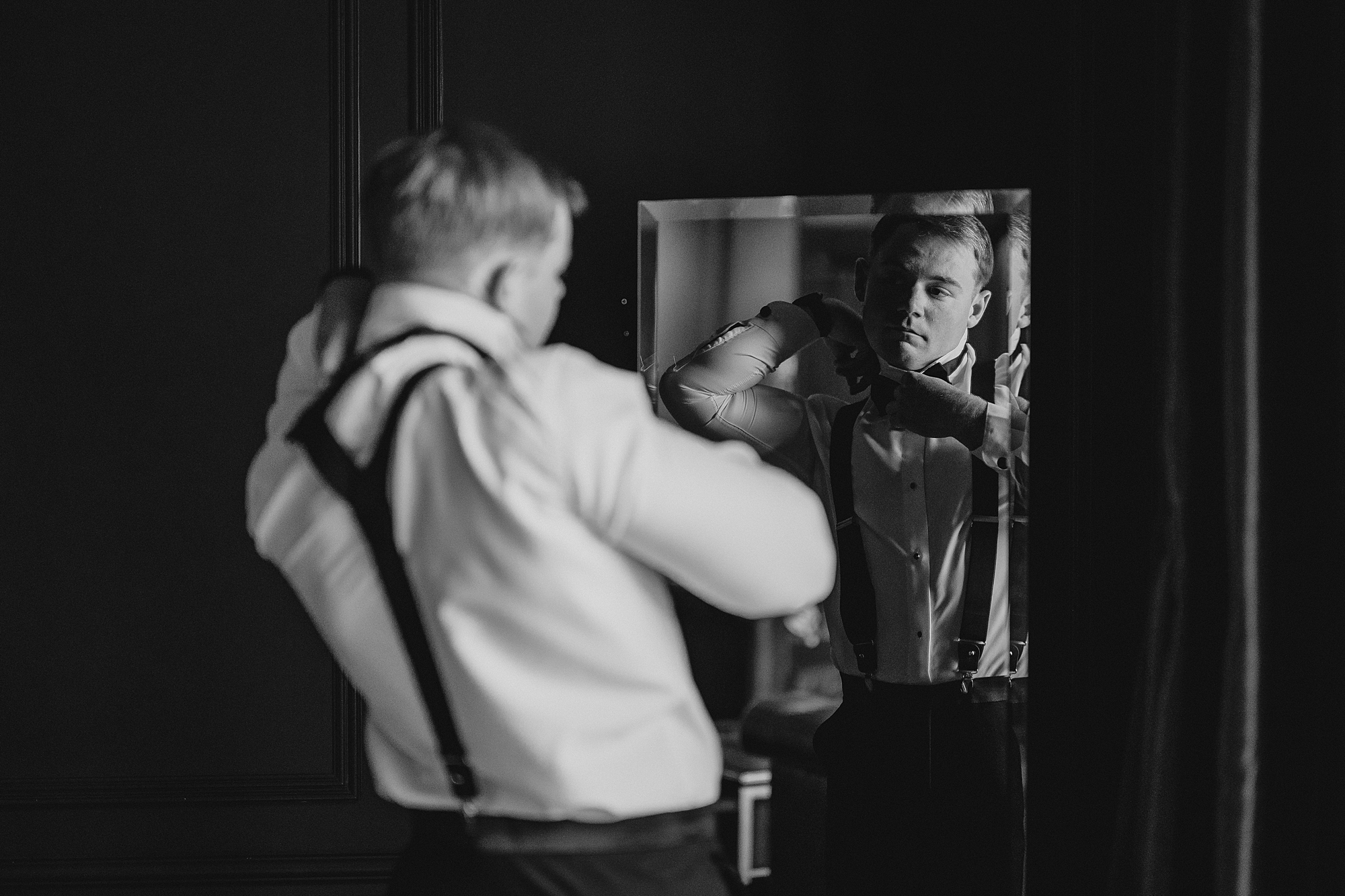 groom adjusts suspenders while looking in mirror 
