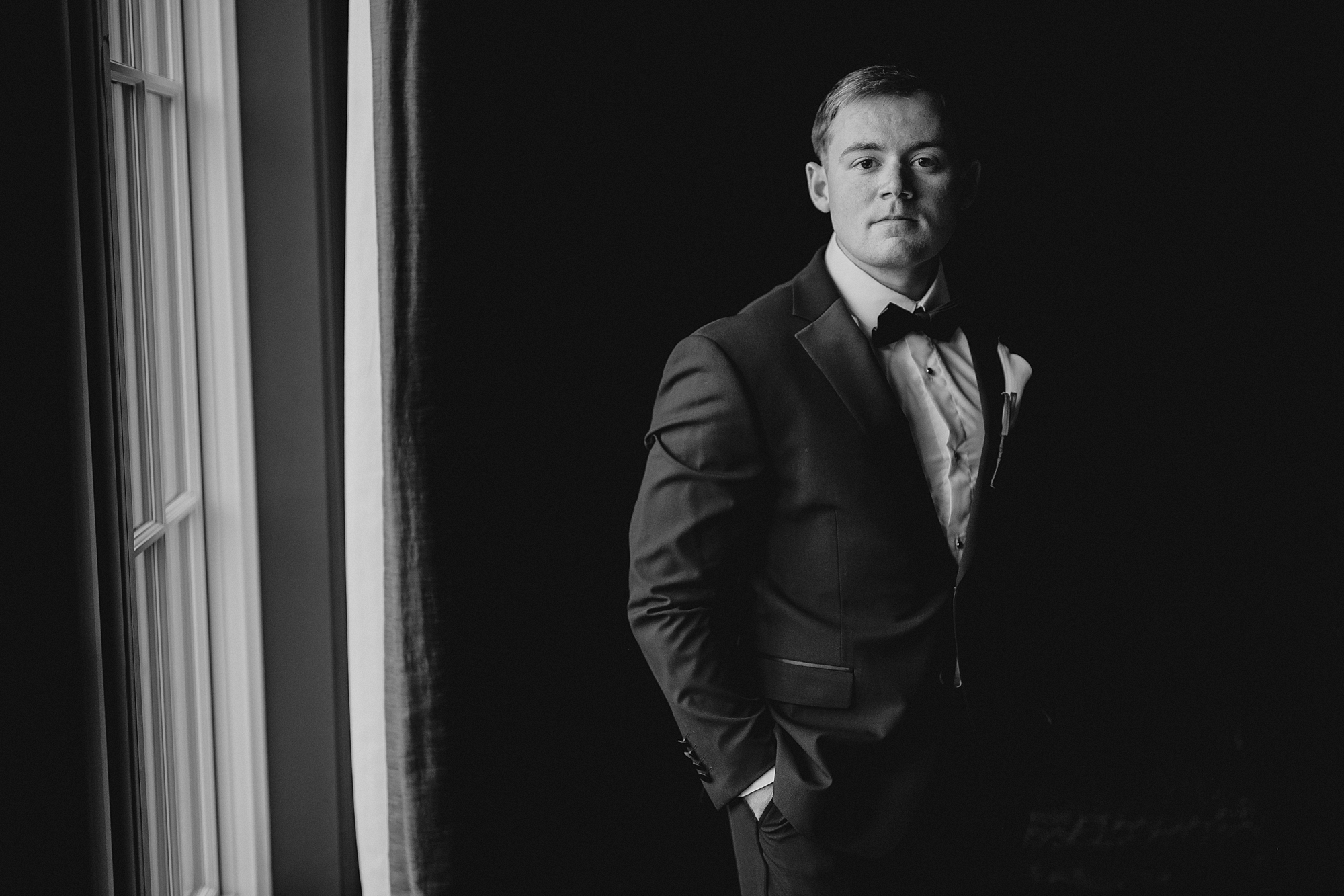 groom stands with hands in pockets of black suit by window in suite at Park Chateau