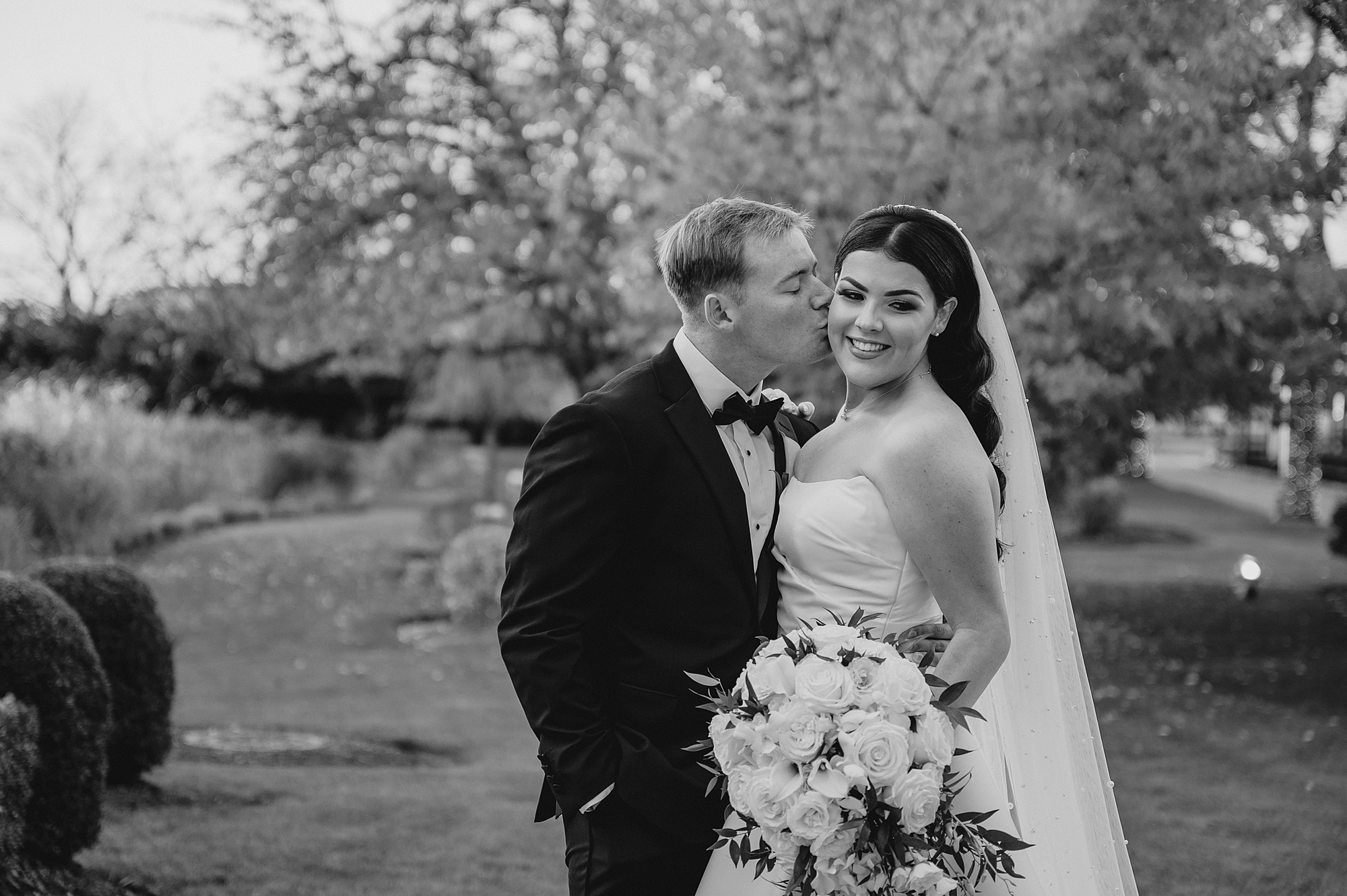 groom kisses bride's cheek hugging her in gardens of Park Chateau