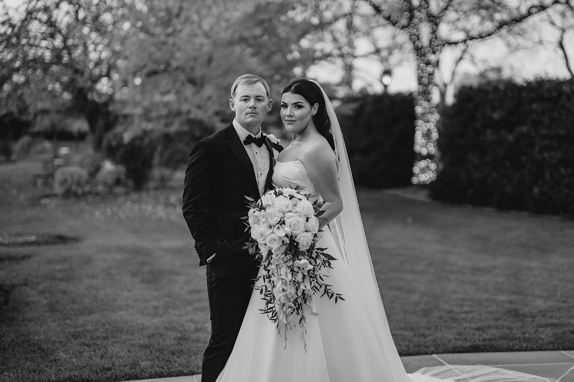 bride and groom stand with straight faces in gardens of Park Chateau
