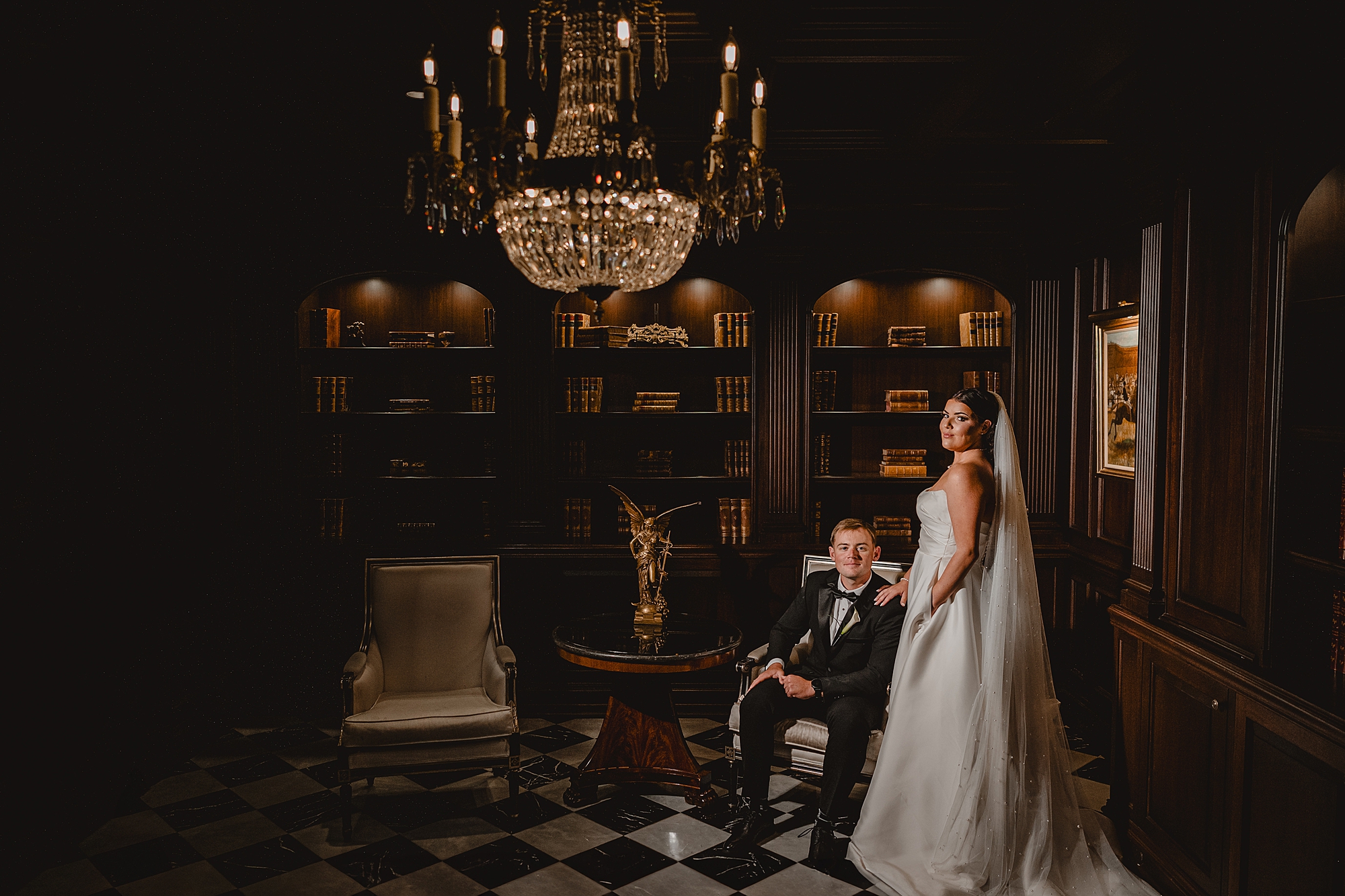 groom sits in chair in library at Park Chateau with bride standing behind him 