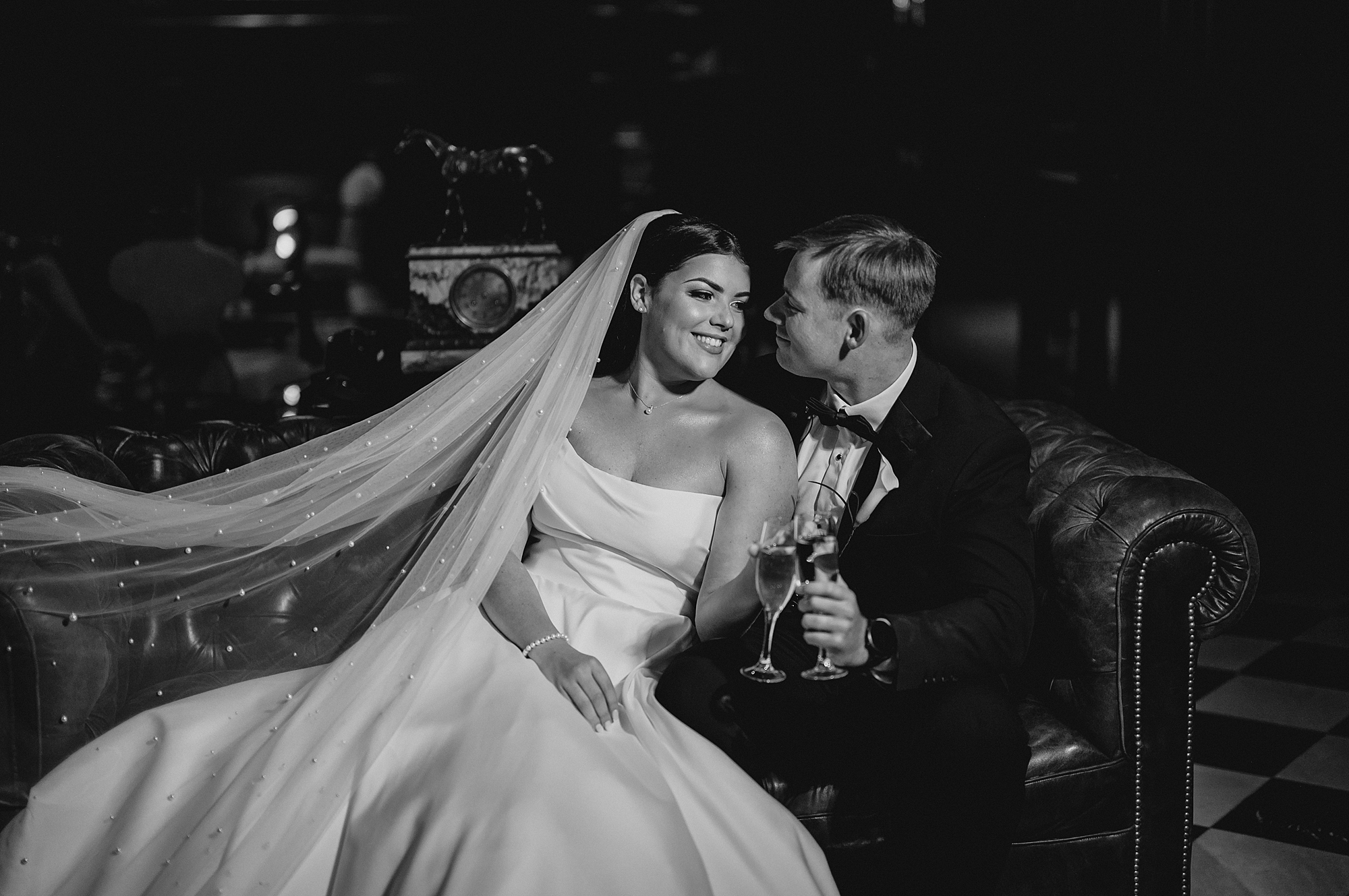bride and groom hug on leather couch toasting glasses of wine 